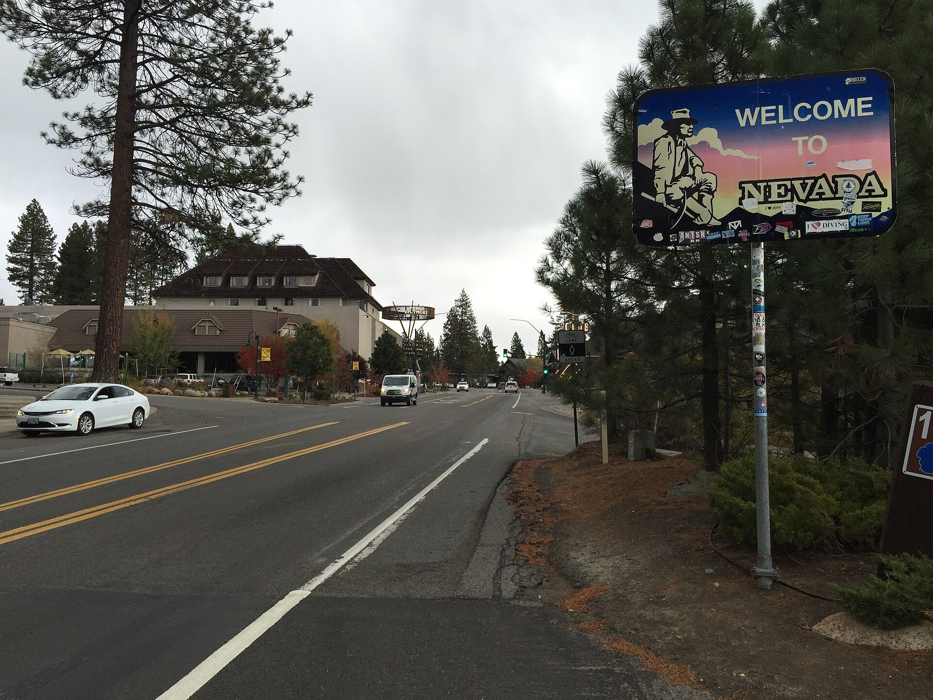 A 'Welcome to Nevada' highway sign beside a road lined with pines and mountain buildings.