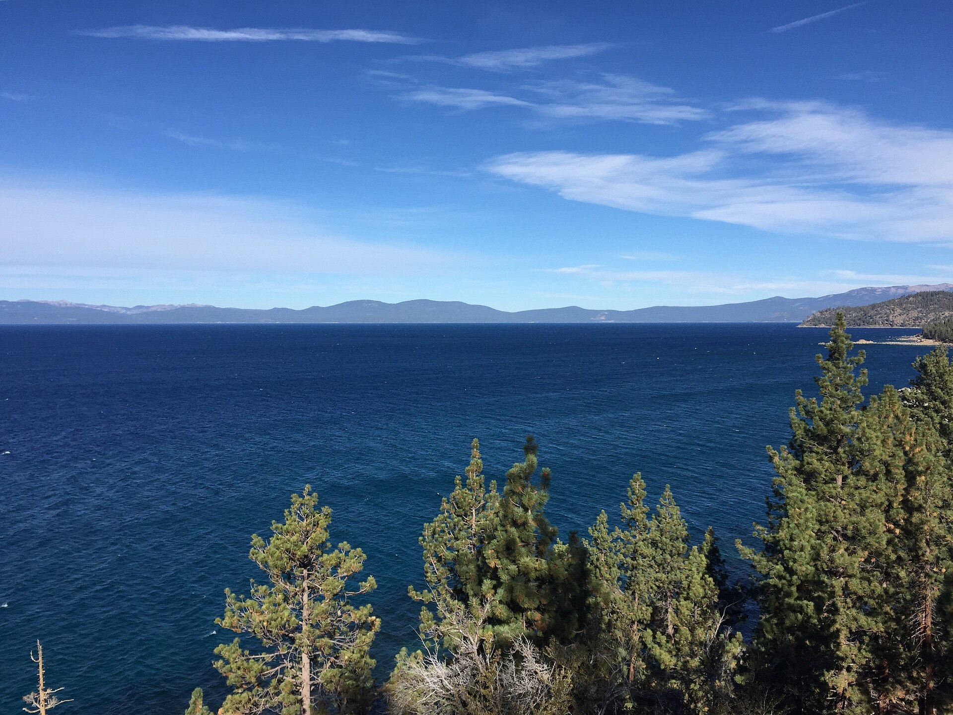 Deep blue lake water under a bright sky, framed by evergreen trees in the foreground.