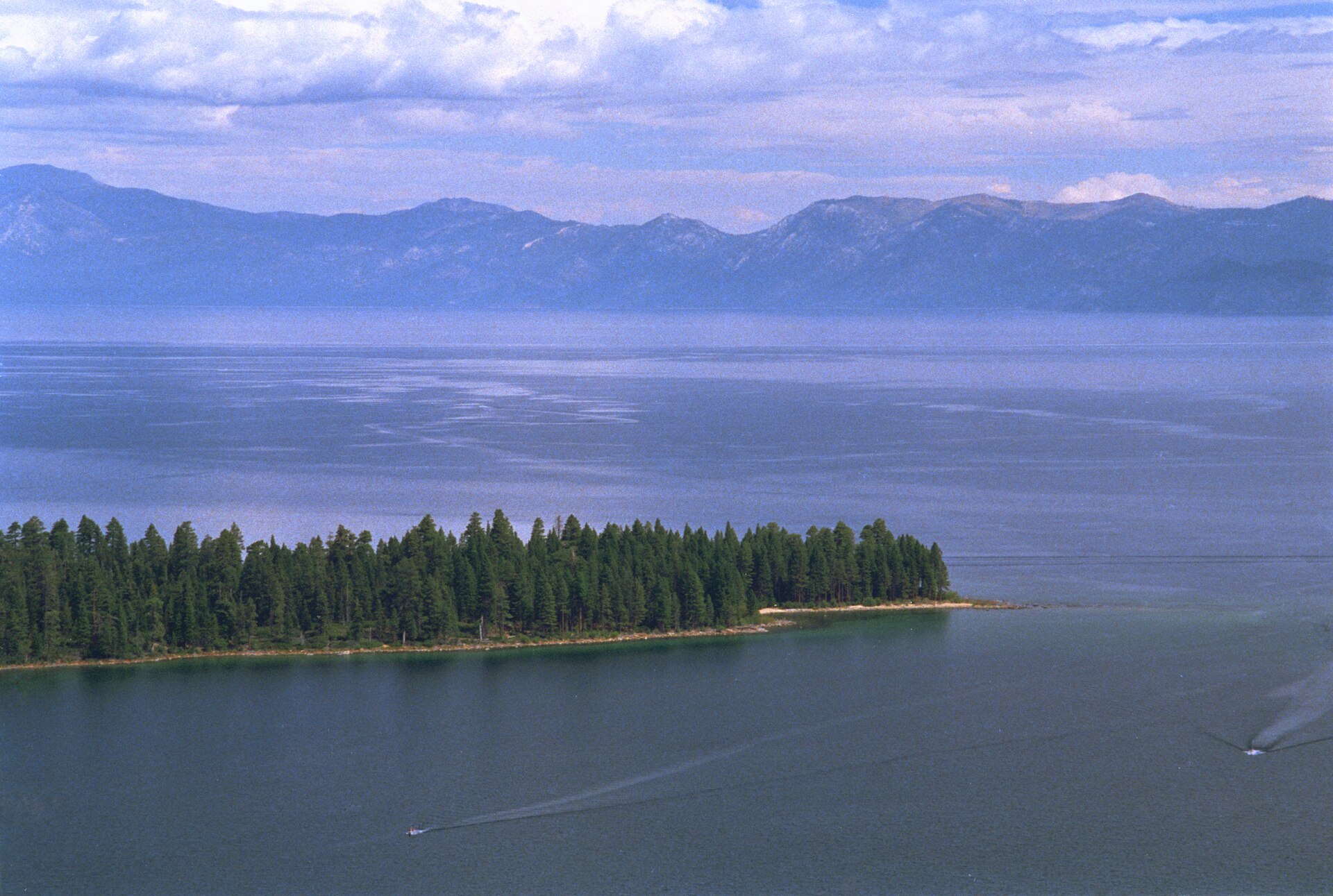 A tree-covered peninsula jutting into calm blue water with hazy mountains across the lake.
