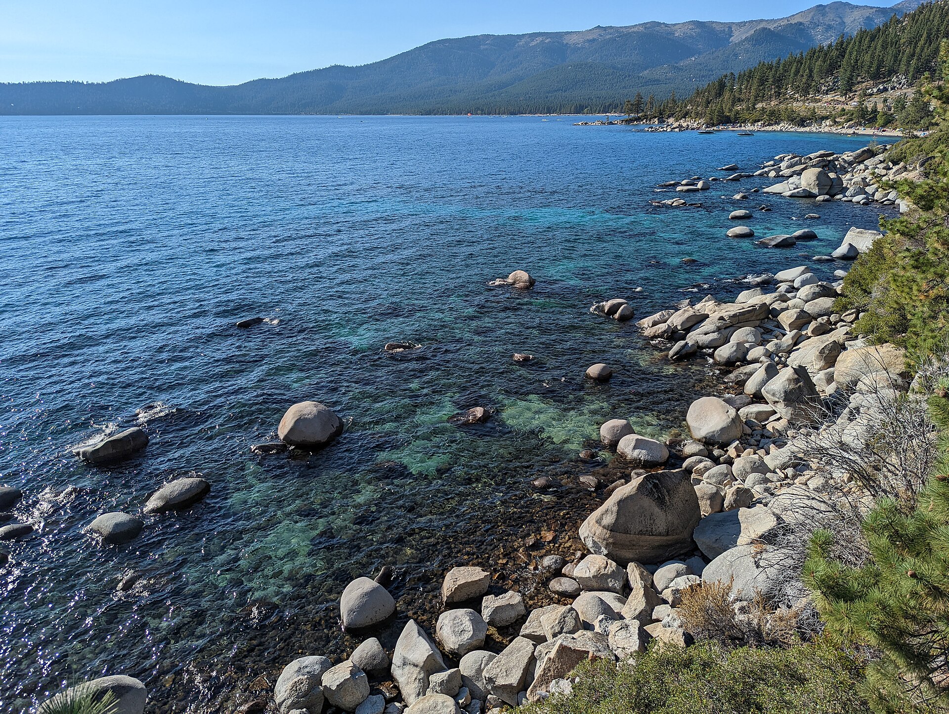 Clear turquoise water lapping boulders on a rocky shore with forested mountains across the lake.