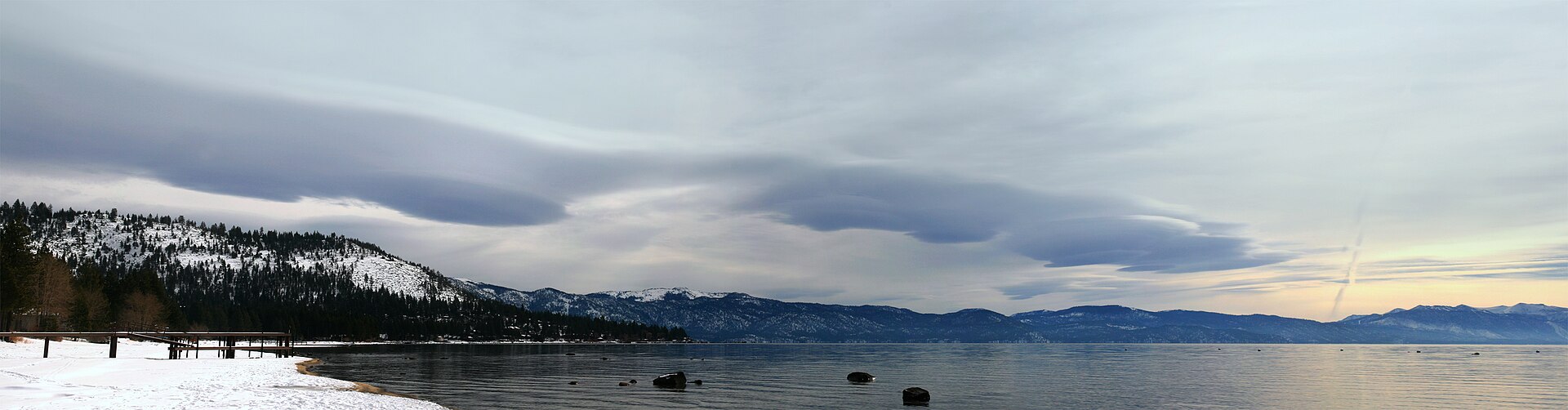 Smooth lens-shaped clouds hovering over snow-dusted mountains and a calm gray lake.