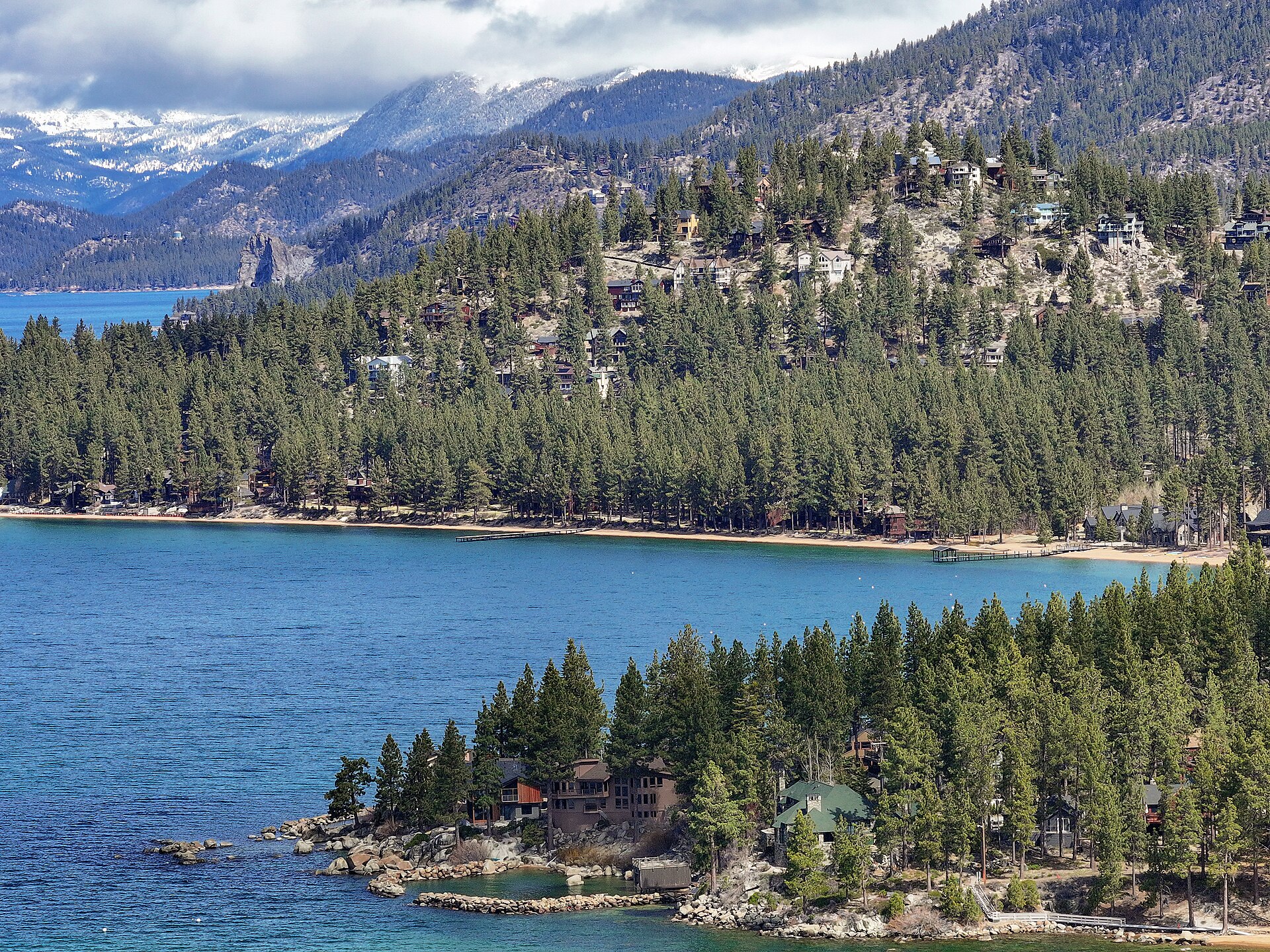 A curving bay of blue water lined with pines and houses, backed by forested mountains.