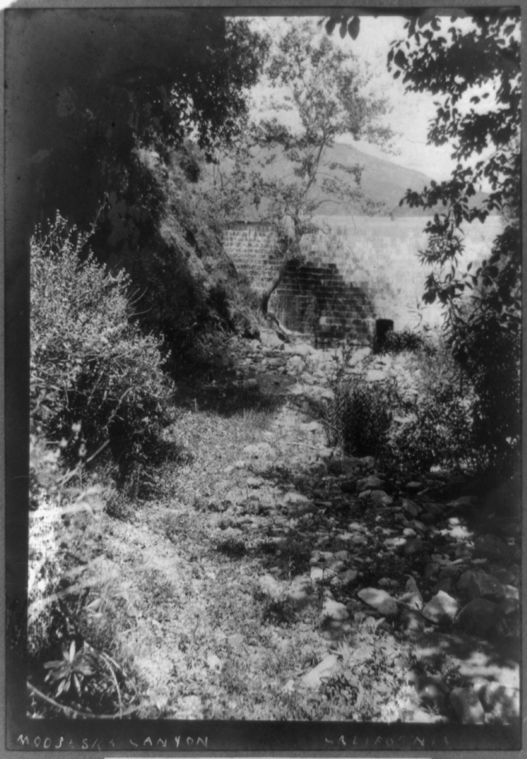 A black-and-white 19th-century view of a rocky creek bed with a stone dam and hills beyond.