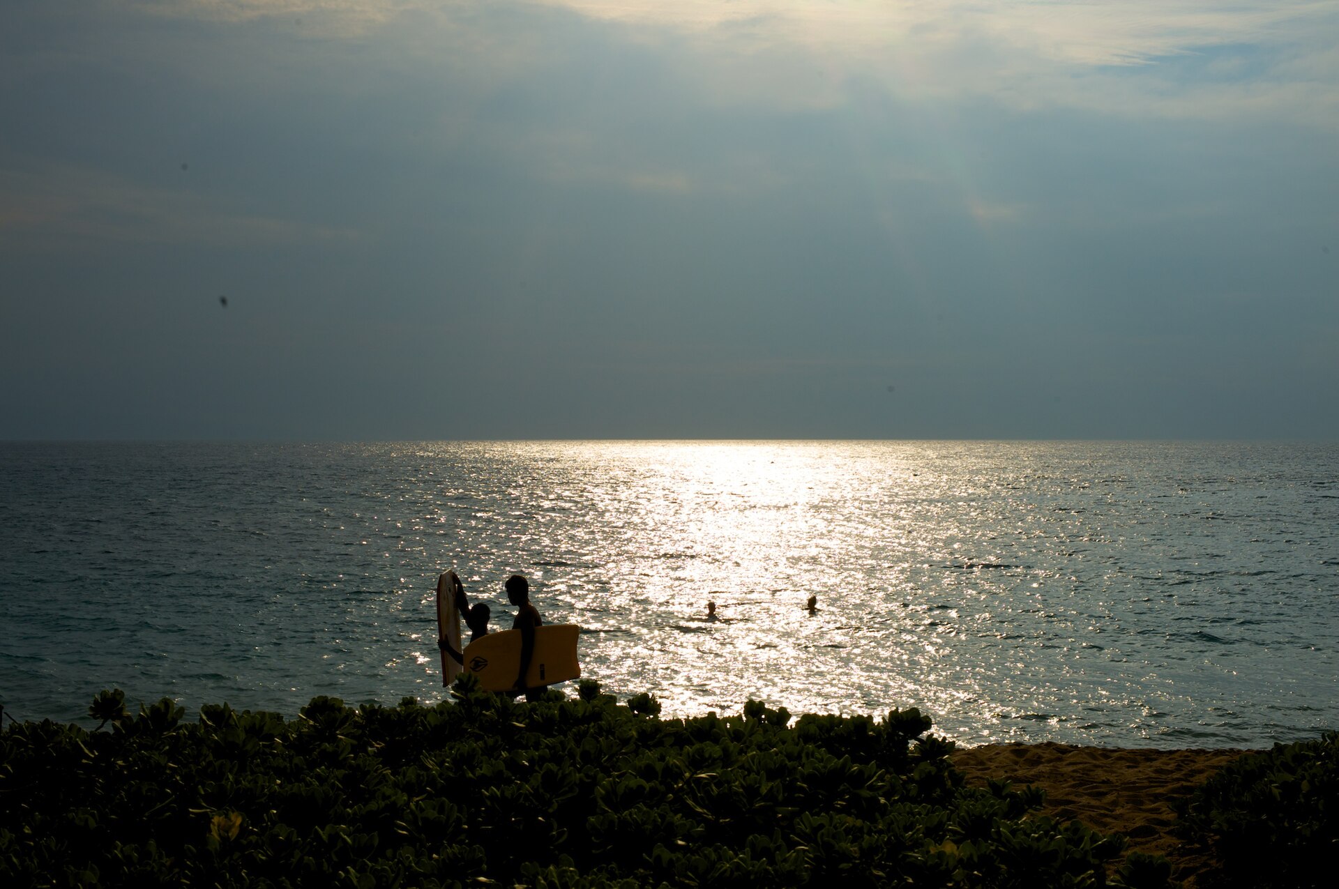 Silhouetted bodyboarders walking into glittering water under a bright hazy sunset.