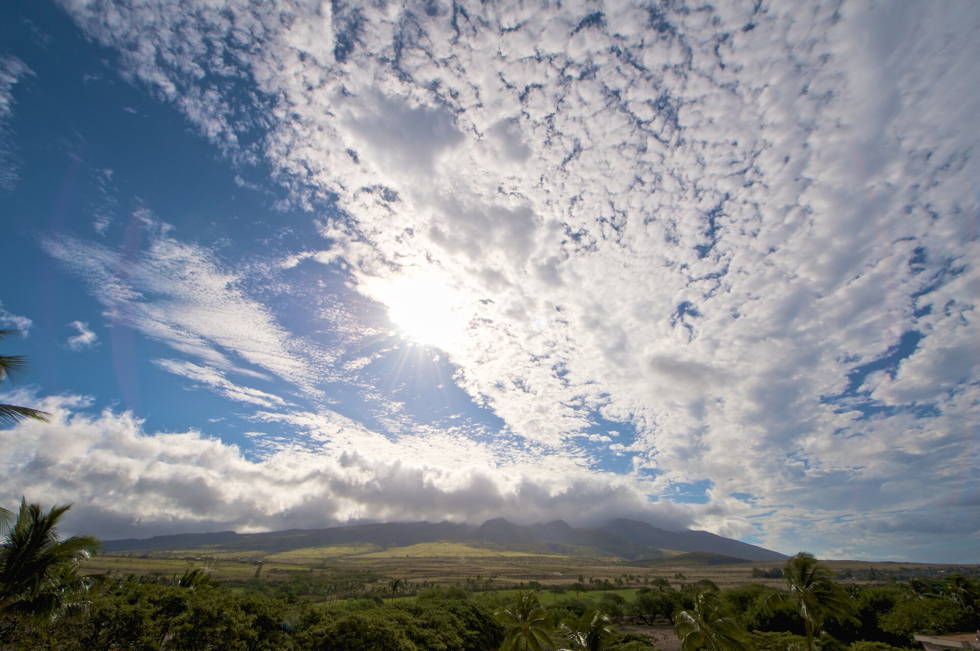 A bright sunburst through rippled clouds above green lowlands and the Mauna Kahalawai ridge.