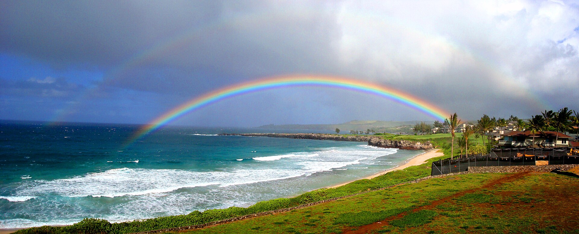 Two rainbows arcing over white surf breaking on a sandy bay with green lawns in the foreground.