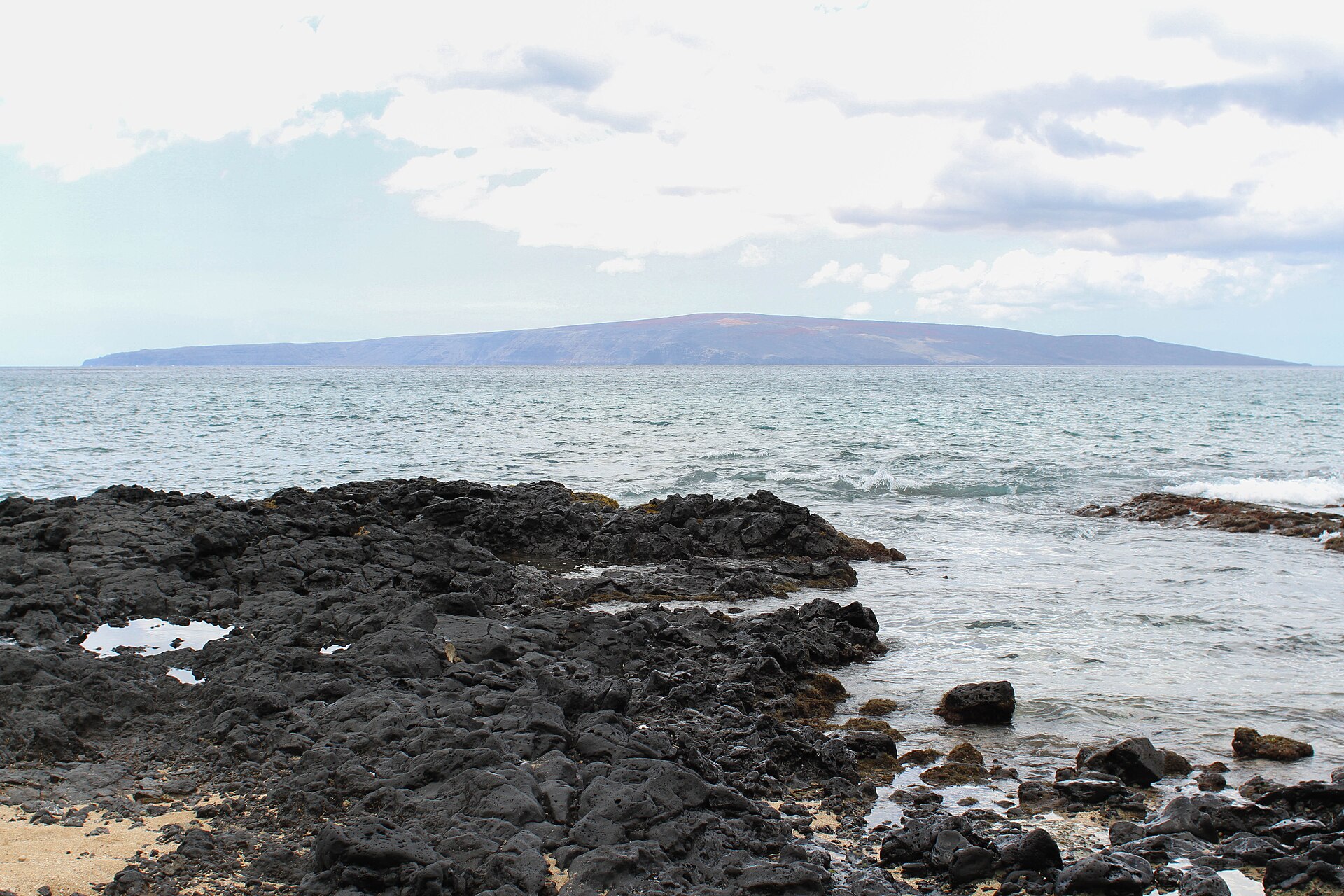 Black lava rocks in the foreground with the flat profile of Kaho'olawe across the water.