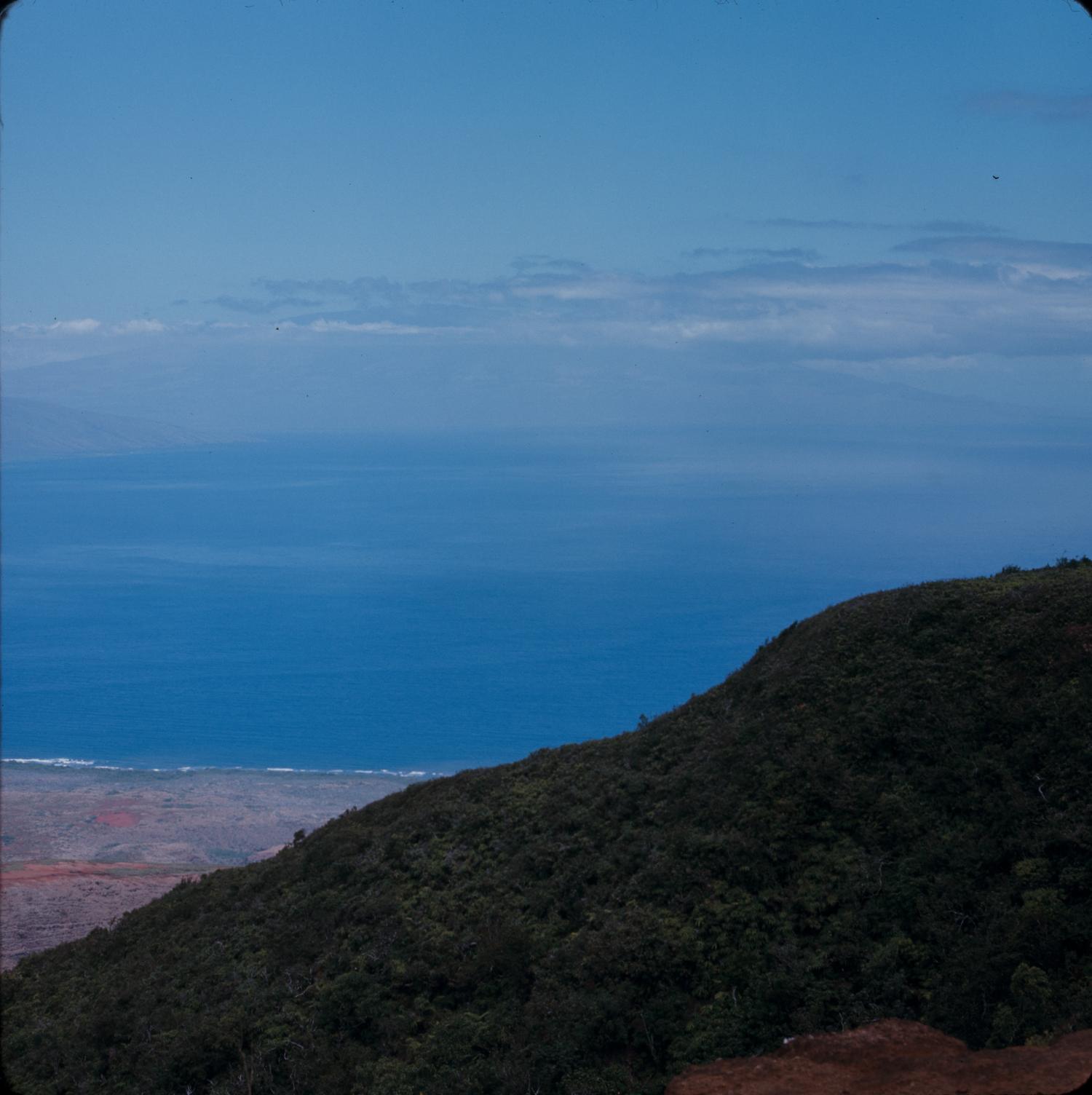 A deep blue ocean channel with the hazy profile of Maui on the horizon, seen from a green hillside on Lana'i.
