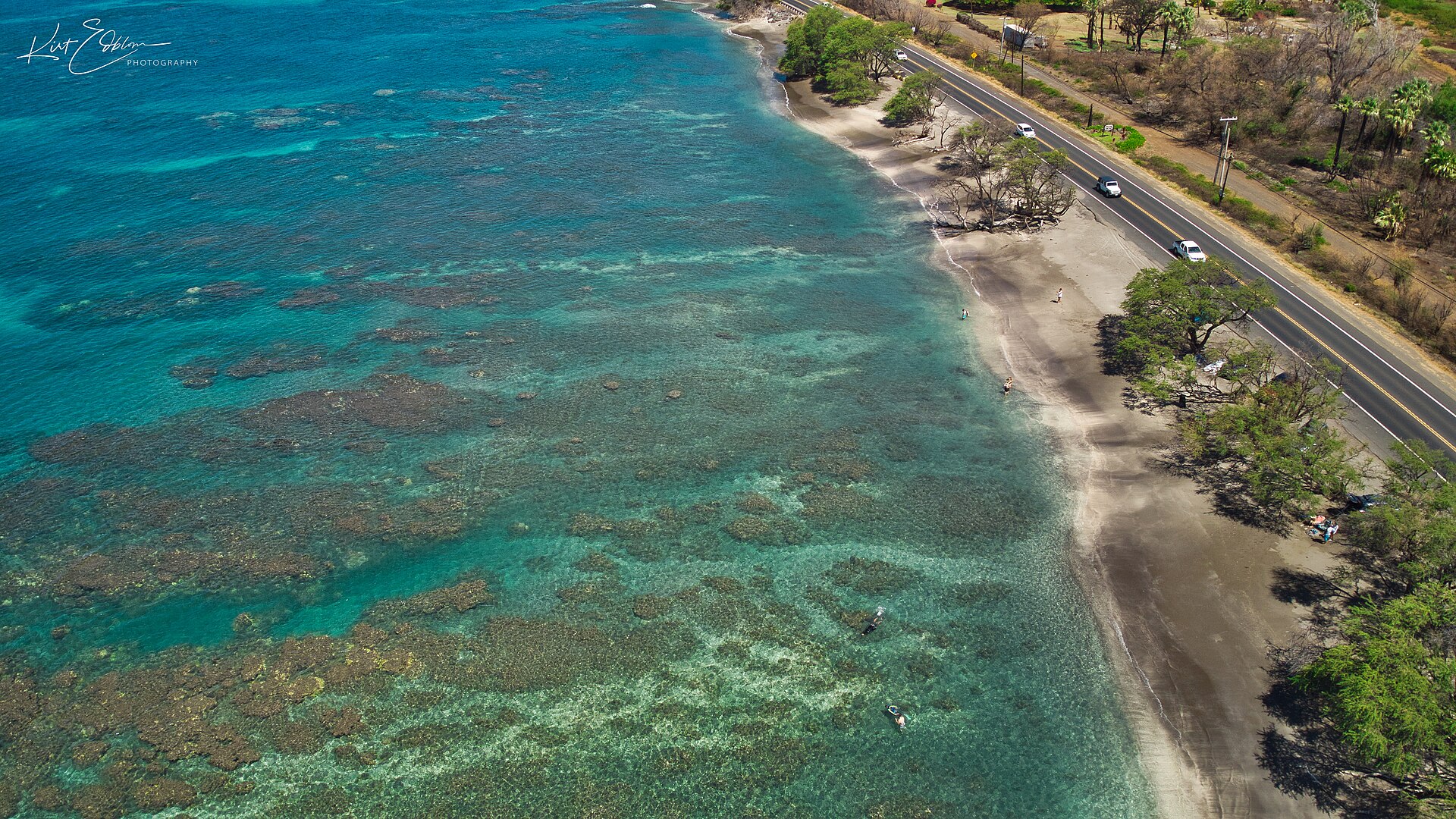 Turquoise water over dark reef patches along a sandy shore with the coastal highway running beside it.