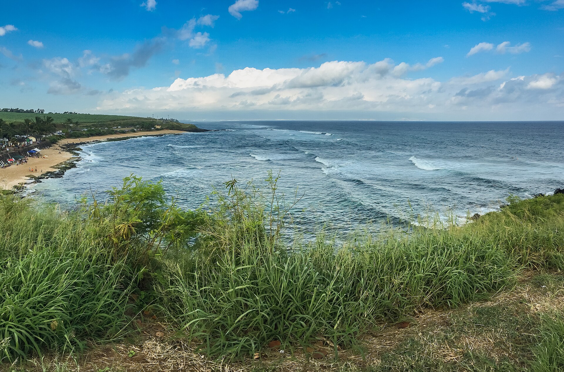 White surf rolling toward a sandy point, seen from a grassy overlook under a bright sky.