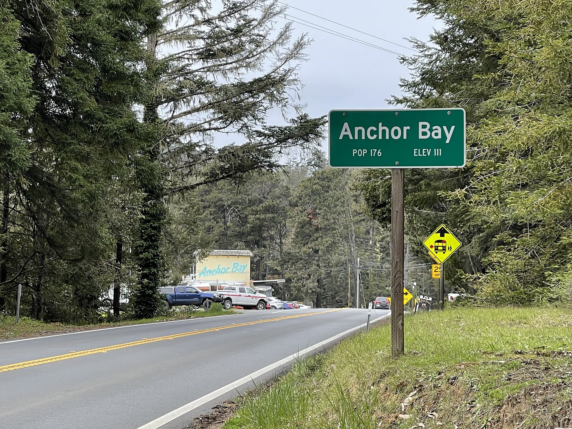 A green highway sign reading 'Anchor Bay, Pop 176, Elev 111' beside a pine-lined road.