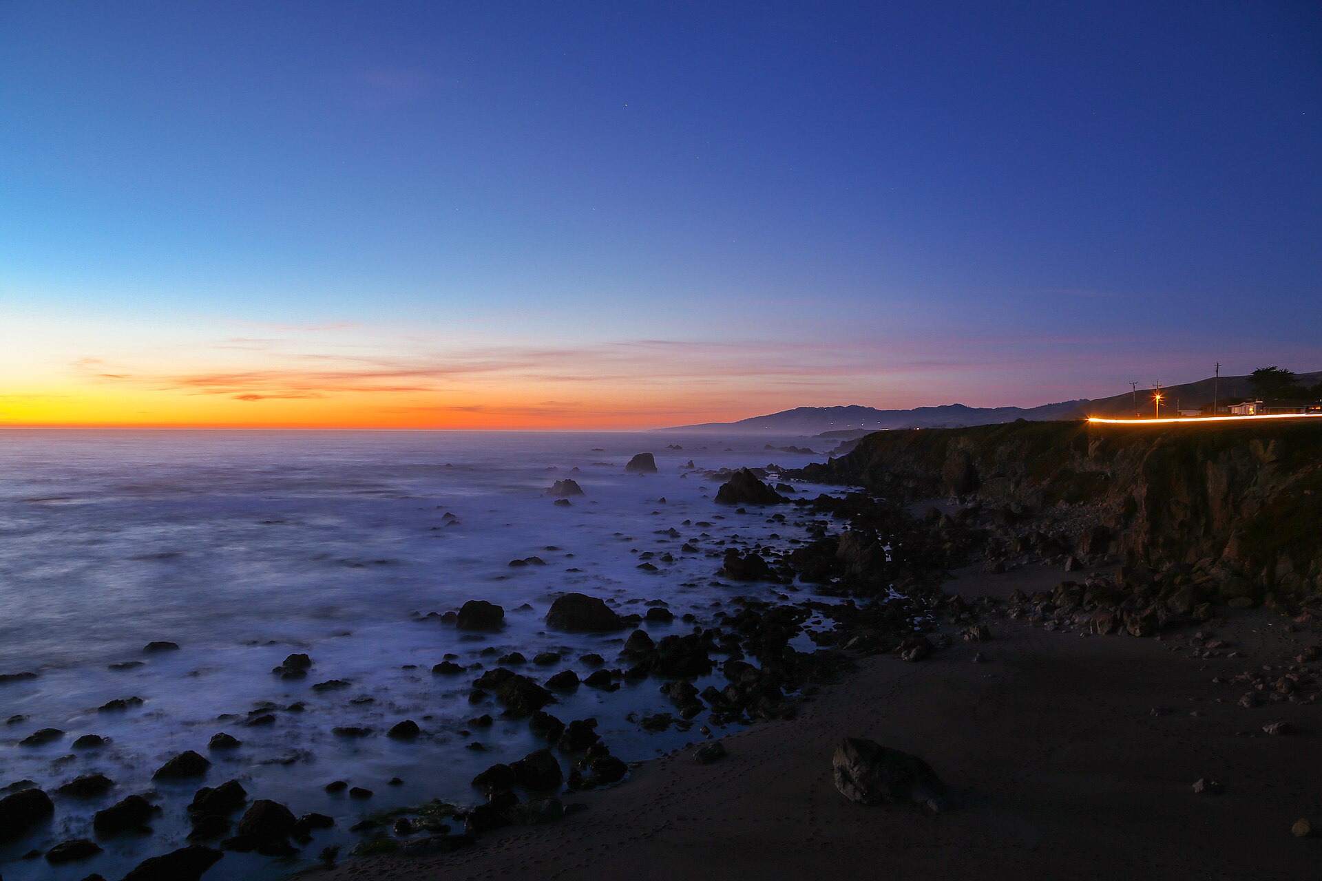 Orange and blue twilight over a rocky shoreline with car lights tracing the cliff road.
