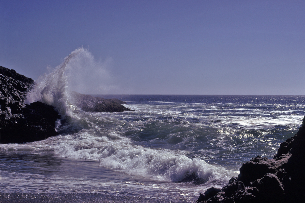 White surf exploding against dark rocks under a clear blue sky.