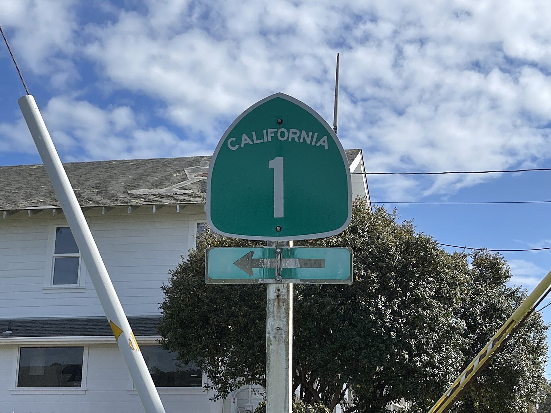A green California Highway 1 shield on a post in front of a white house.