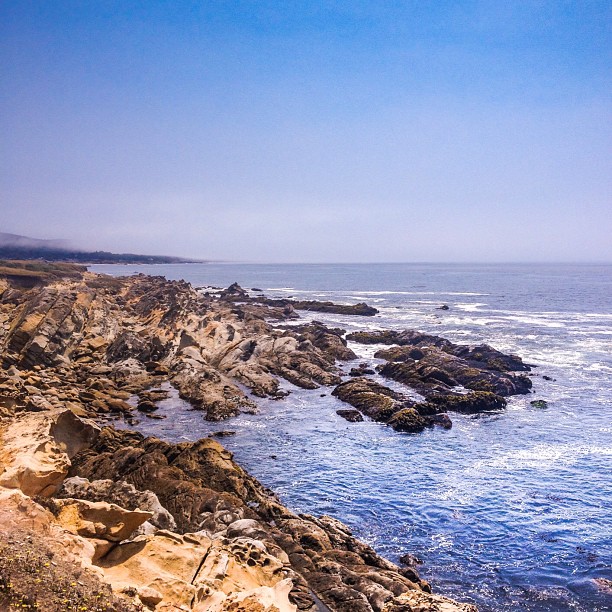 Jagged rock formations and tide pools stretching along a blue ocean under a hazy sky.