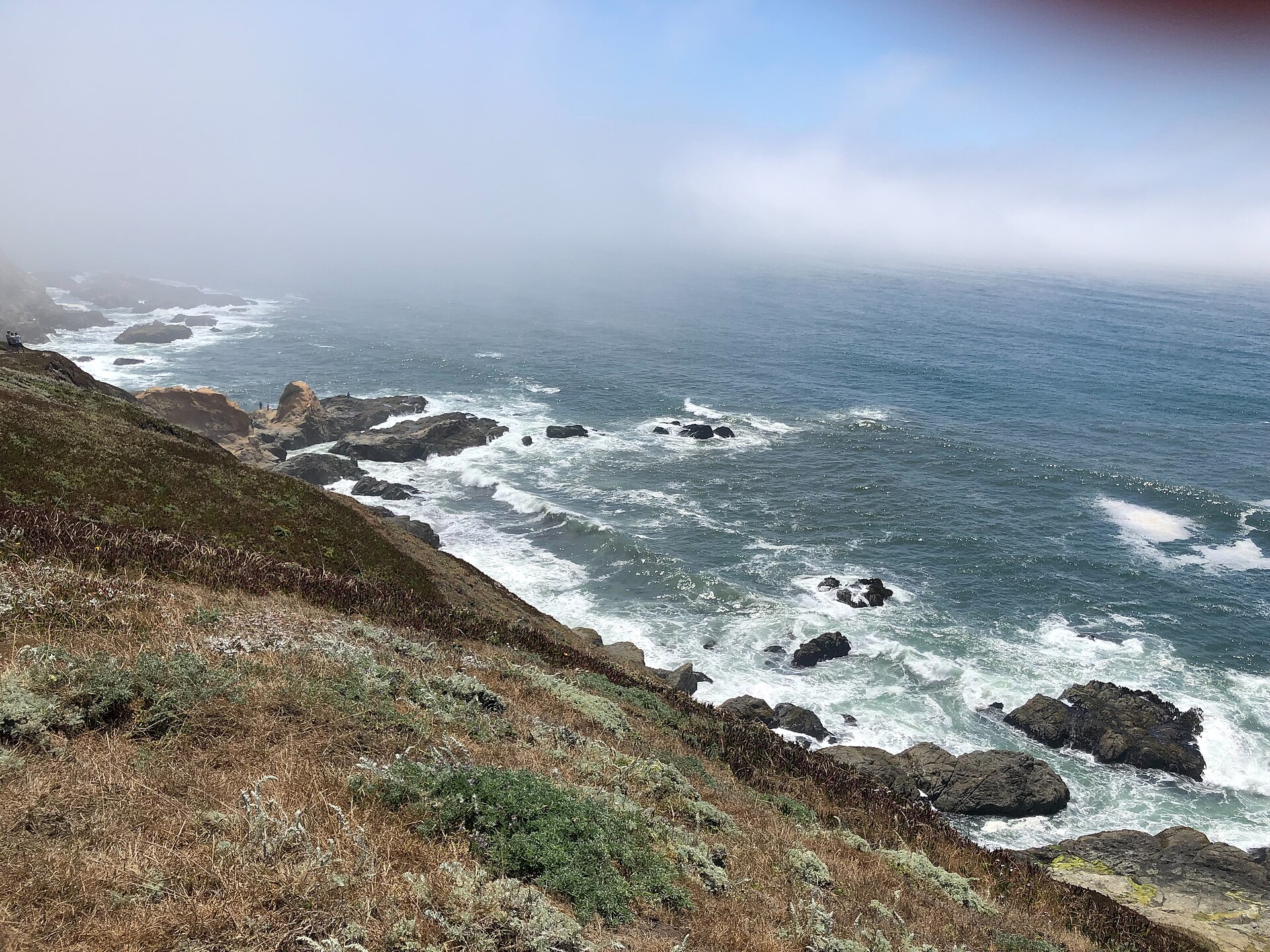 Waves washing over a rocky point with coastal hills fading into ocean mist.
