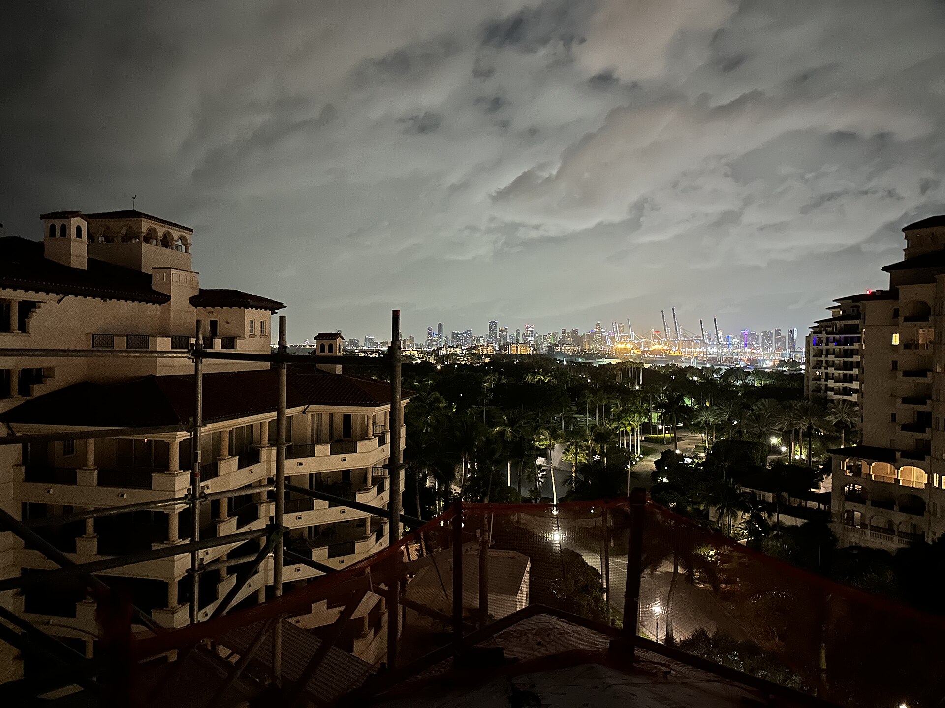 City lights and construction cranes glowing across dark water, framed by island buildings.