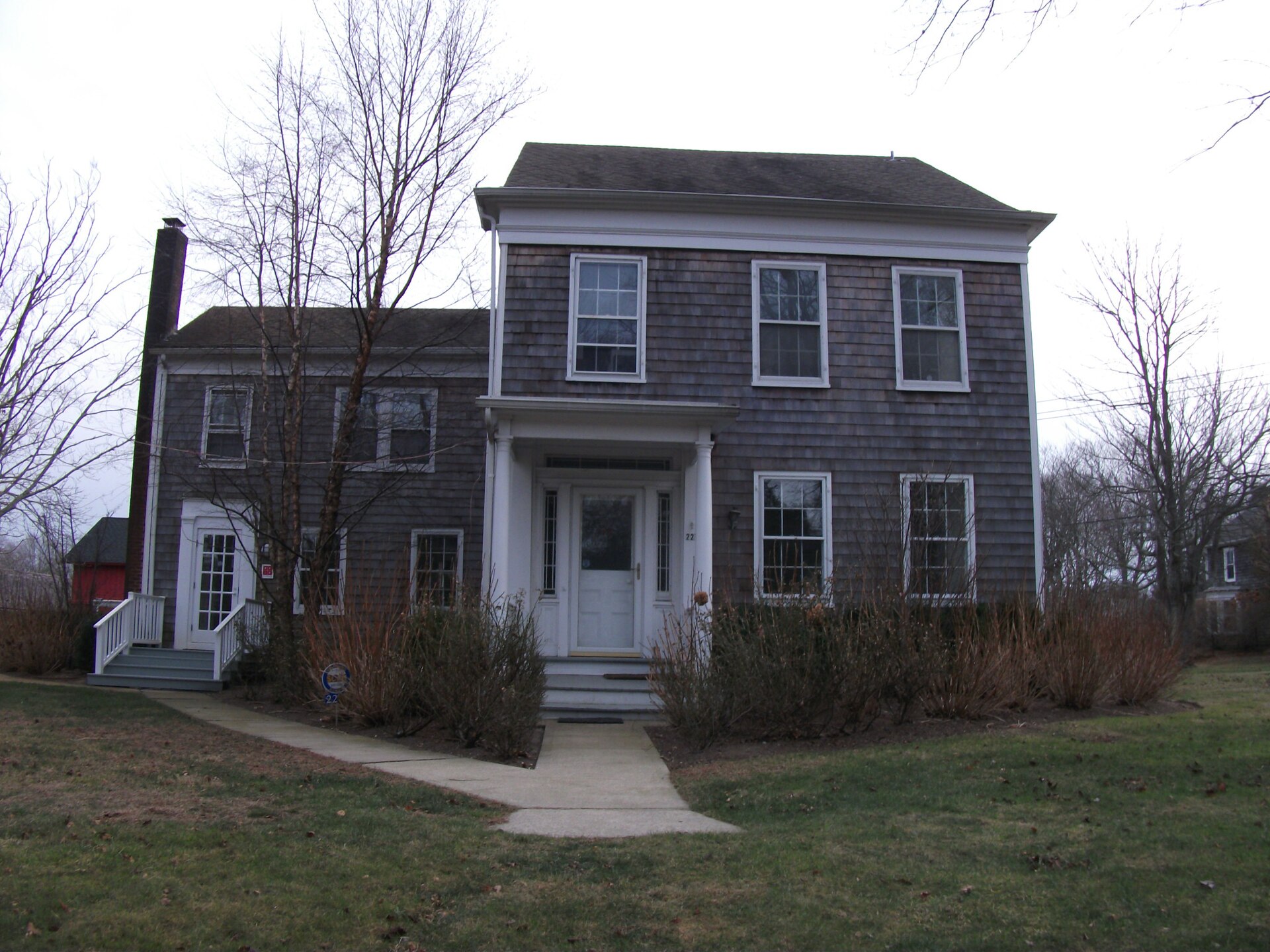A gray-shingled colonial house with white trim and a brick chimney on a lawn.