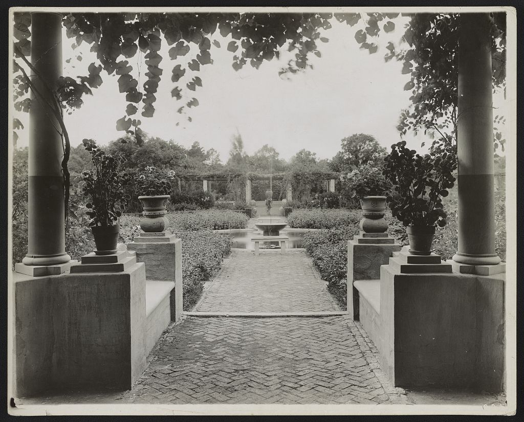 A black-and-white historic view of a formal brick garden with columns, urns, and a central fountain.