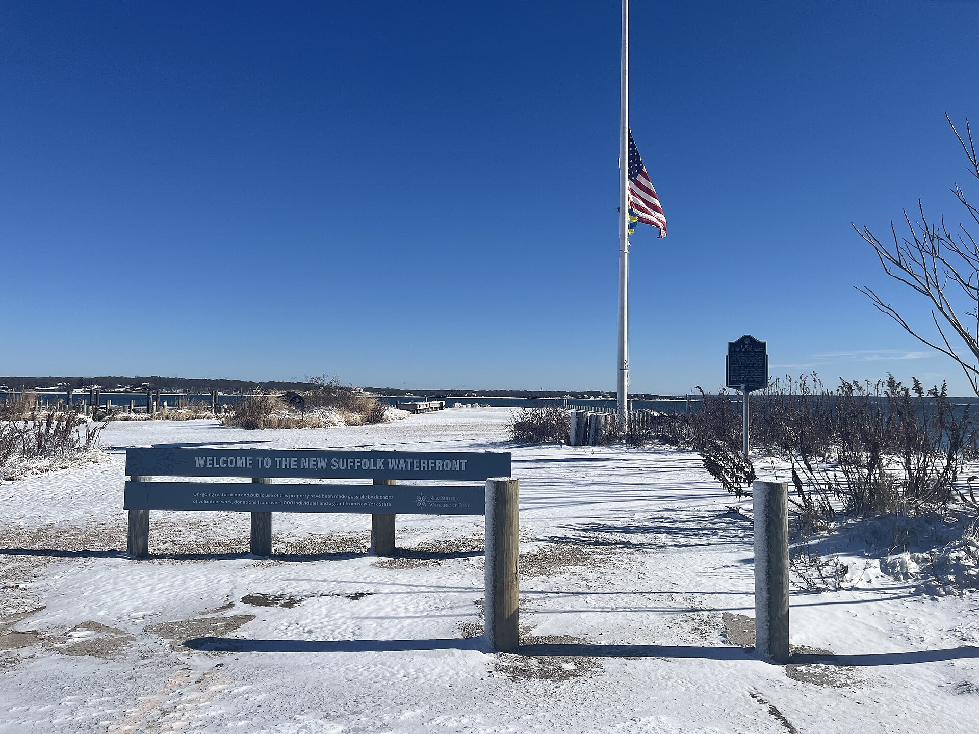 A 'Welcome to the New Suffolk Waterfront' bench on a snowy beach with an American flag.