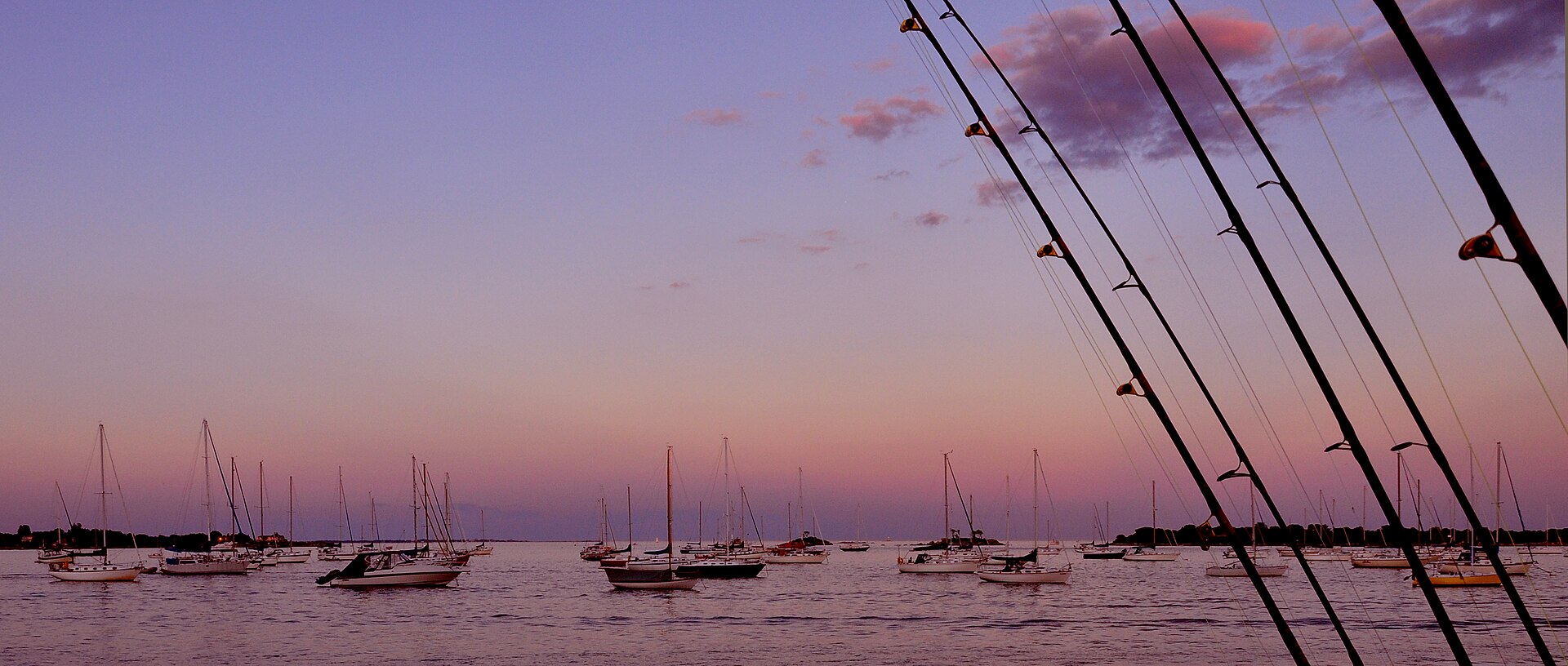 Sailboats moored in Noank's harbor at sunset, with fishing rods in the foreground.