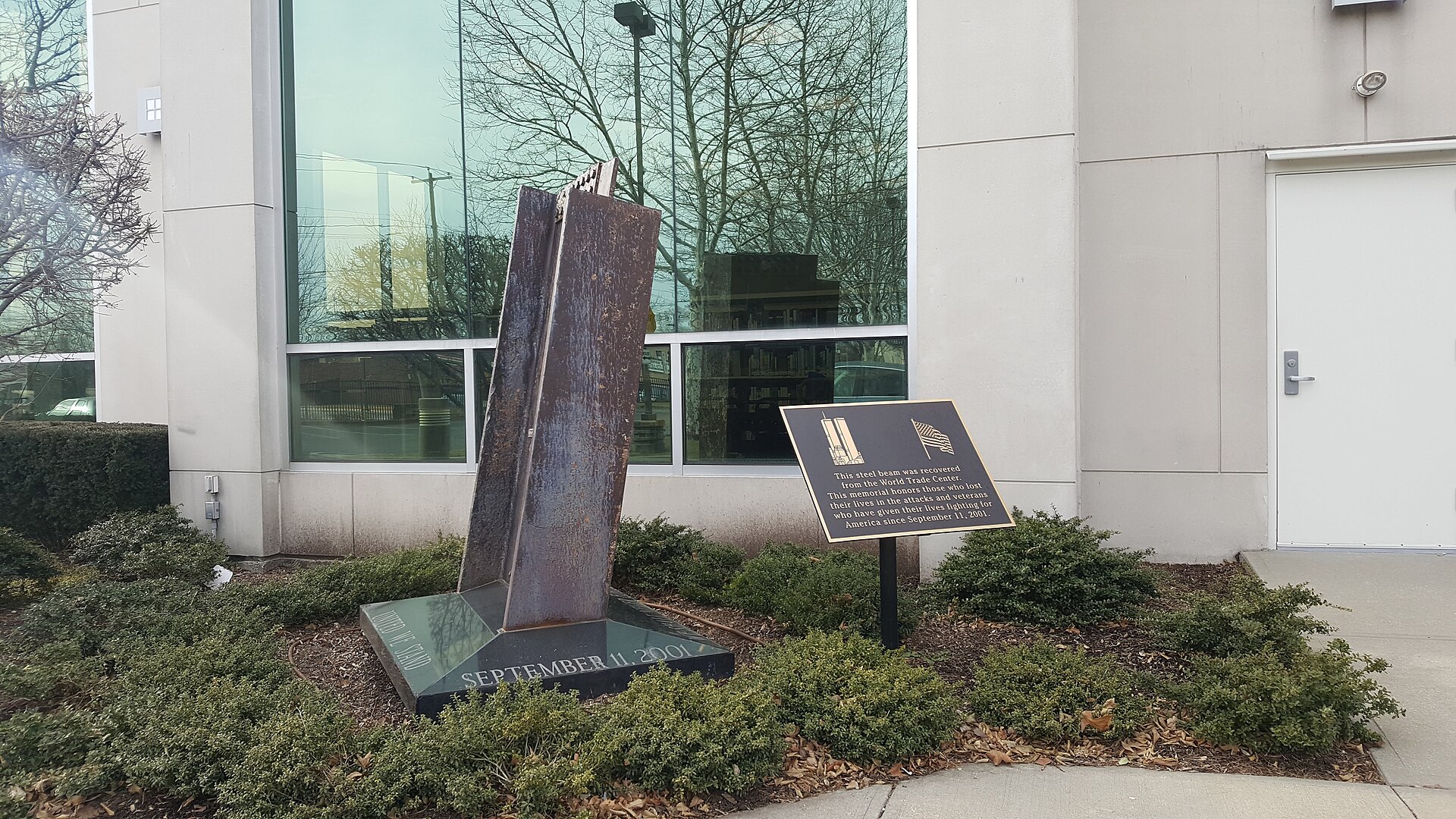 A steel memorial sculpture and plaque in a landscaped bed outside a library building.