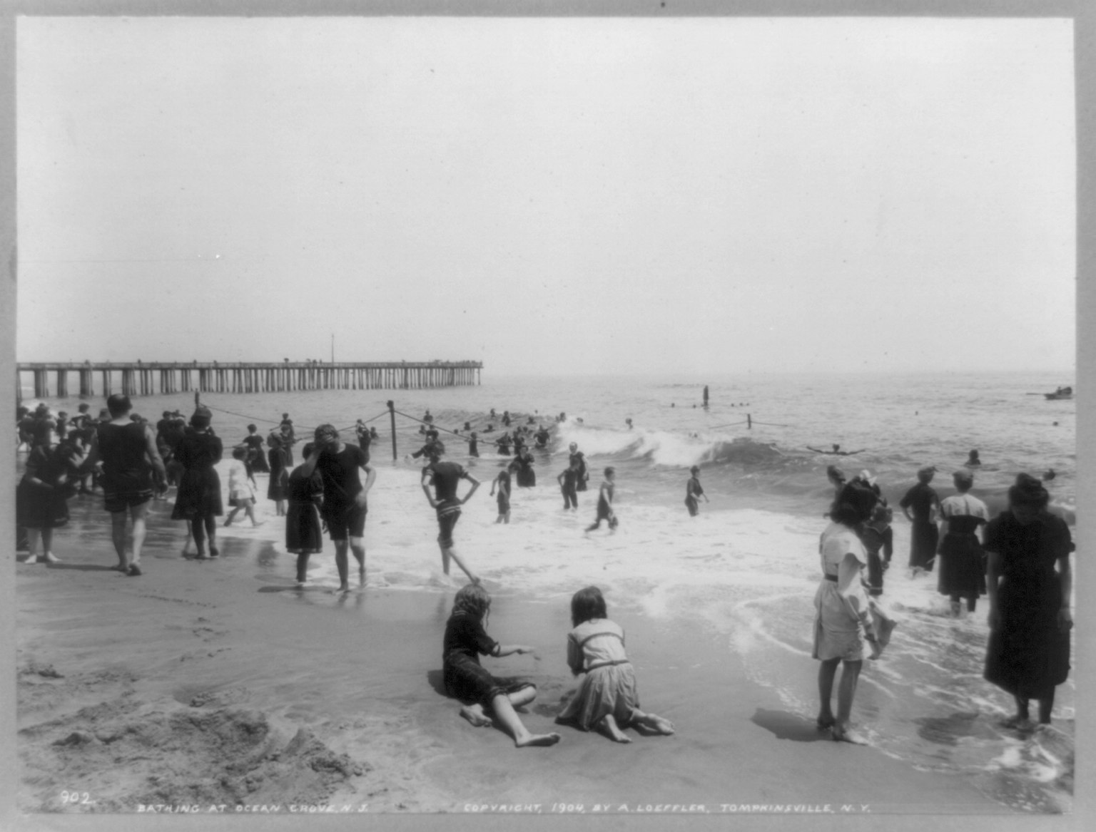 Black-and-white historic photo of crowds bathing at Ocean Grove beach with a pier in the background.