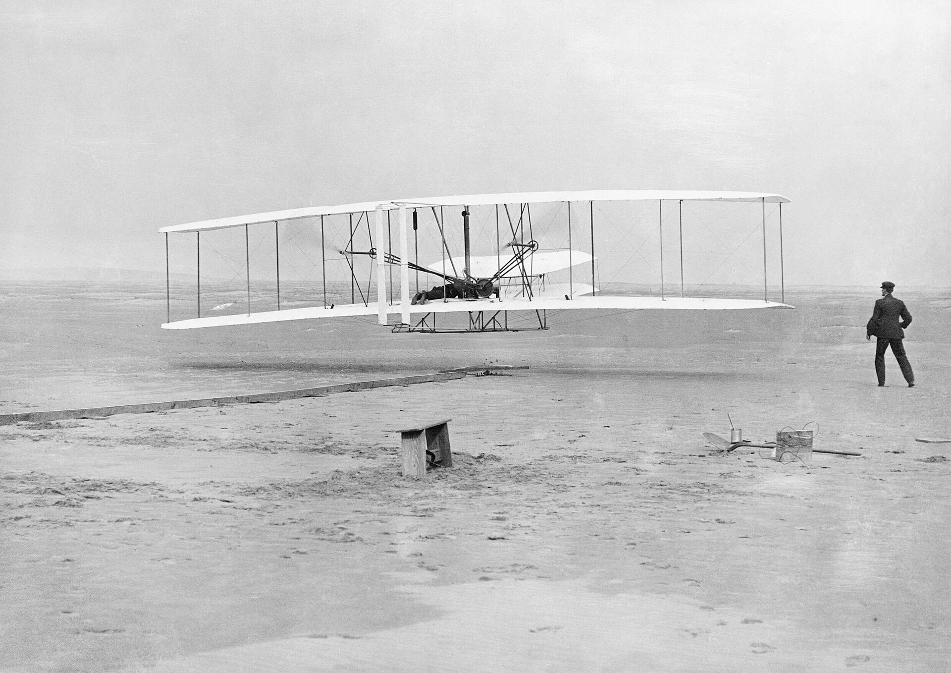 Black-and-white photo of the Wright Flyer airborne over sand with a man running alongside.