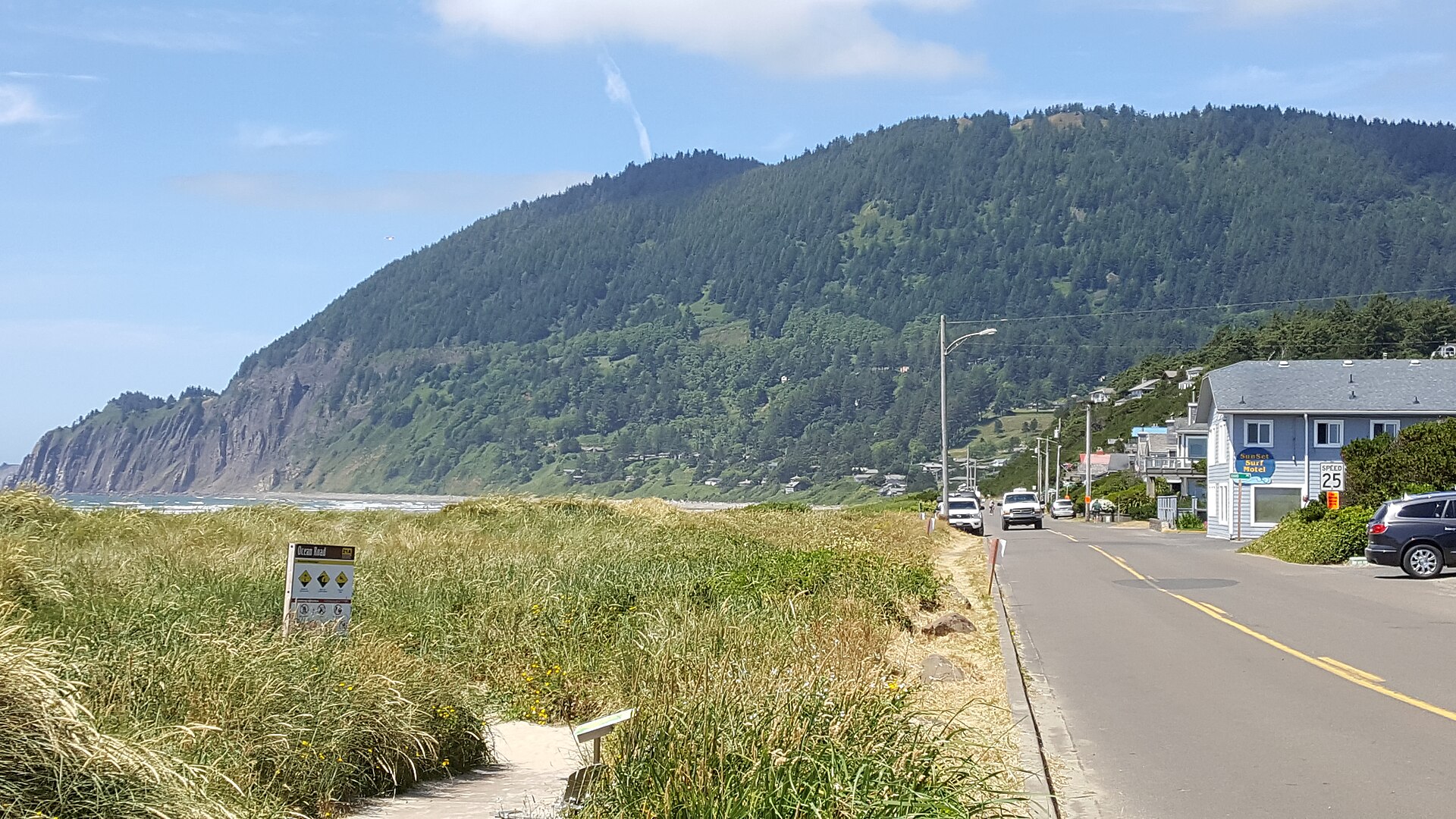 A paved road in Manzanita, Oregon, with grassy dunes on one side and a forested mountain ahead.