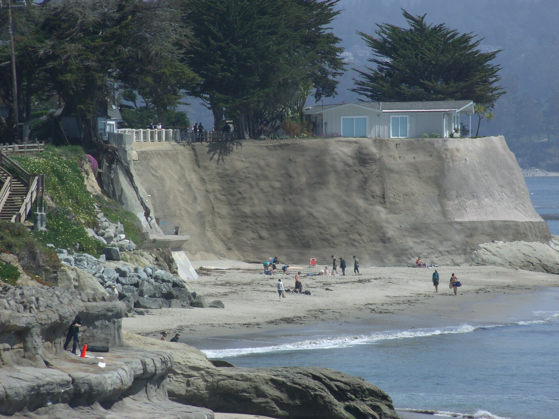Beachgoers on the sand at Pleasure Point below a steep bluff topped with a beach house and cypress trees.