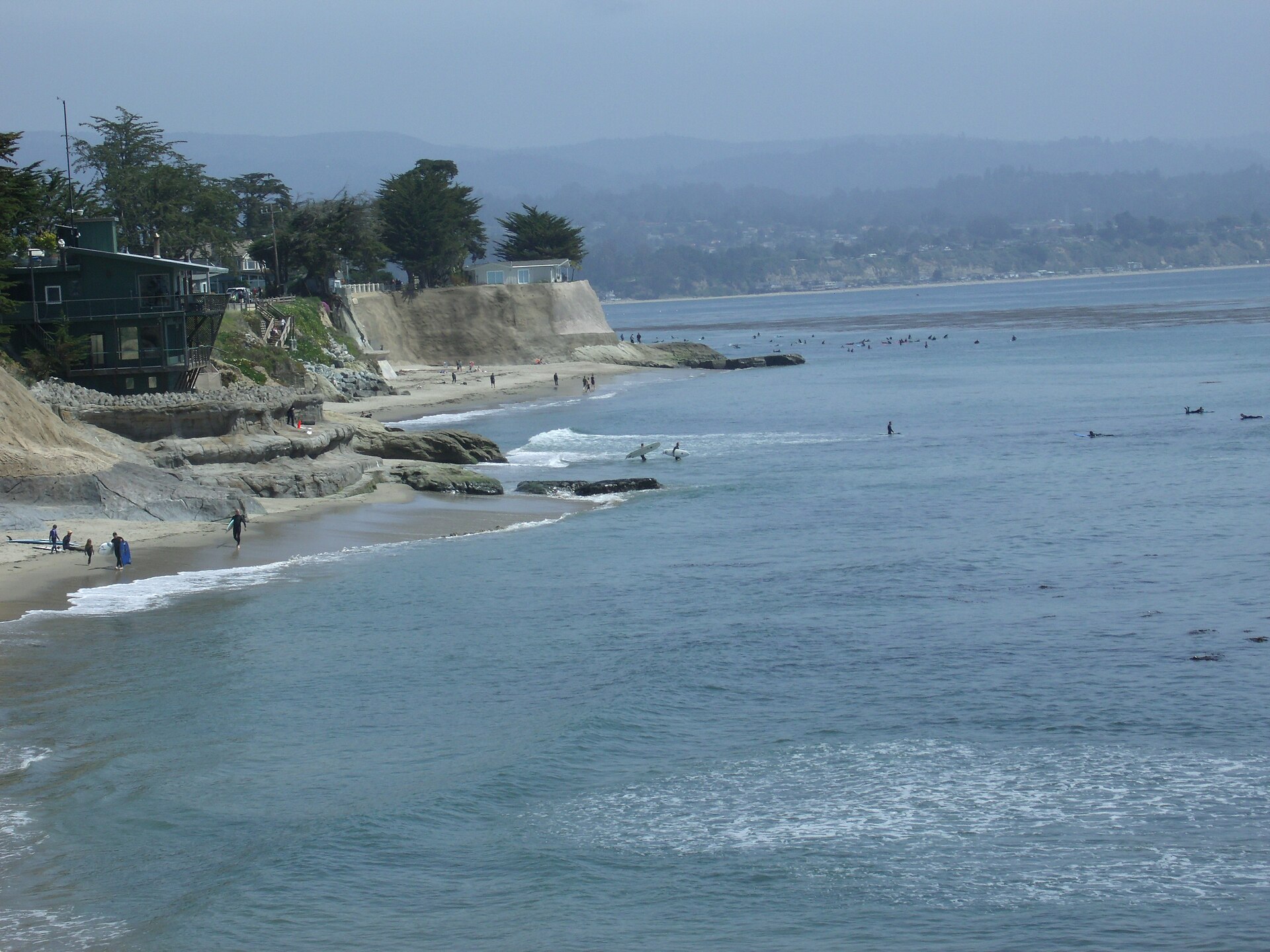 Surfers in the water off Pleasure Point with the rocky shoreline and beach houses on the bluffs.