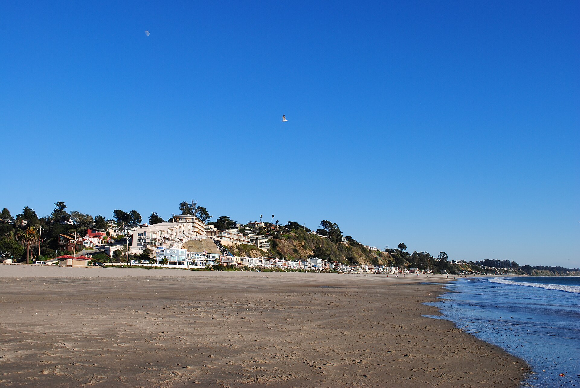A wide sandy beach at Rio Del Mar stretching toward bluff-top homes under a clear blue sky.