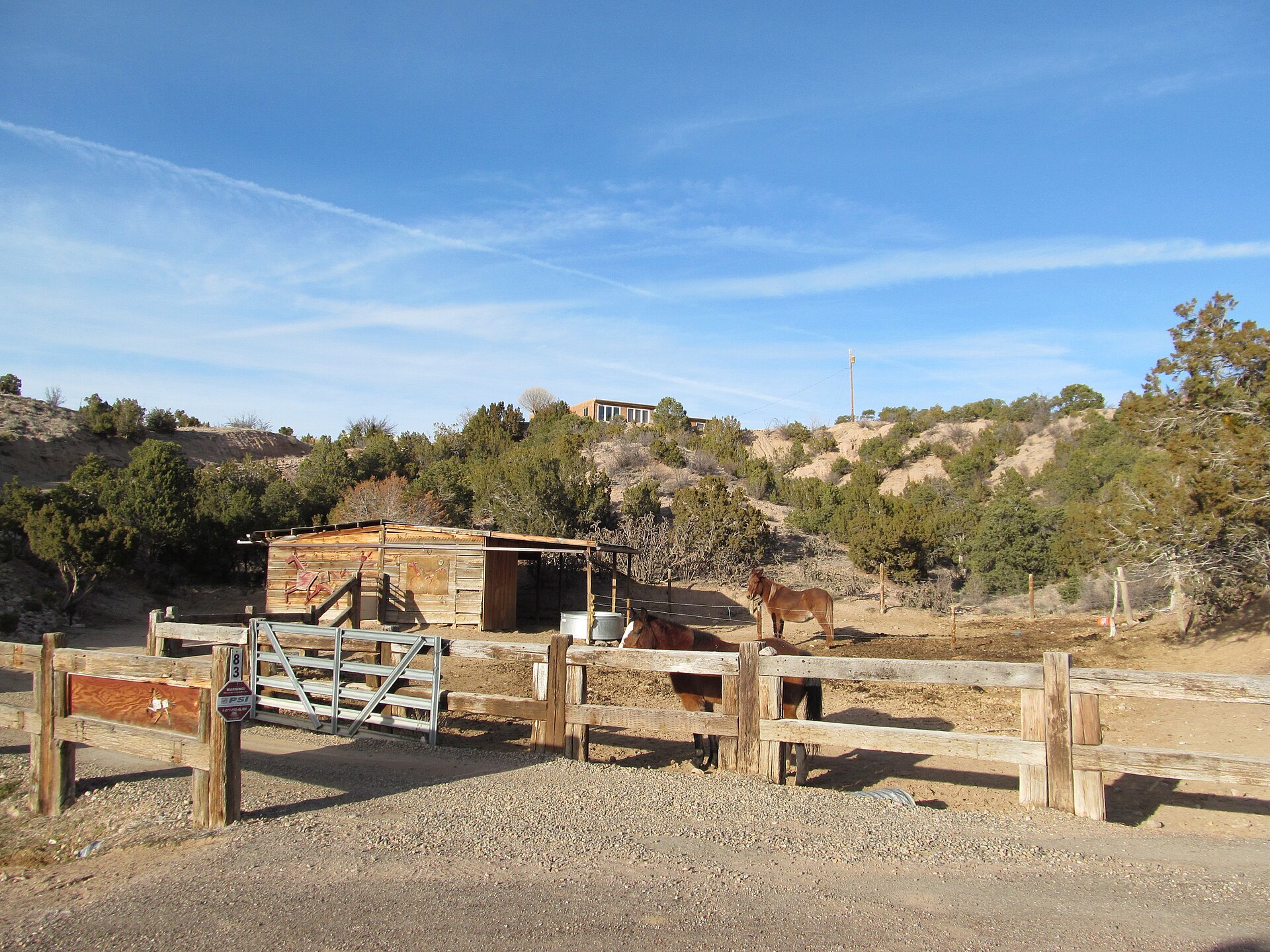 Horses in a fenced corral with a wooden shelter in Chupadero, with pinon-covered hills behind.