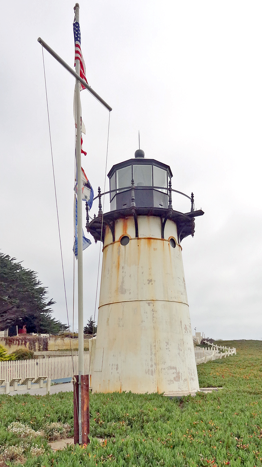 The Point Montara Lighthouse, a white cylindrical tower with a black lantern room, standing on a grassy bluff with an American flag.