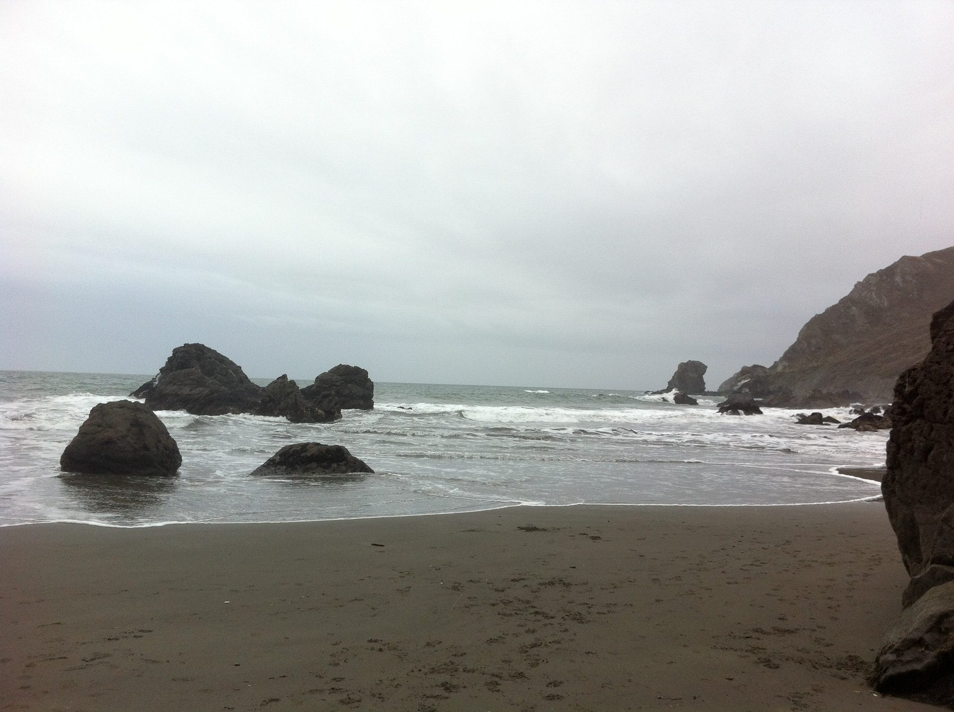 Dark rocks and surf at Muir Beach under overcast skies, with sea stacks offshore.