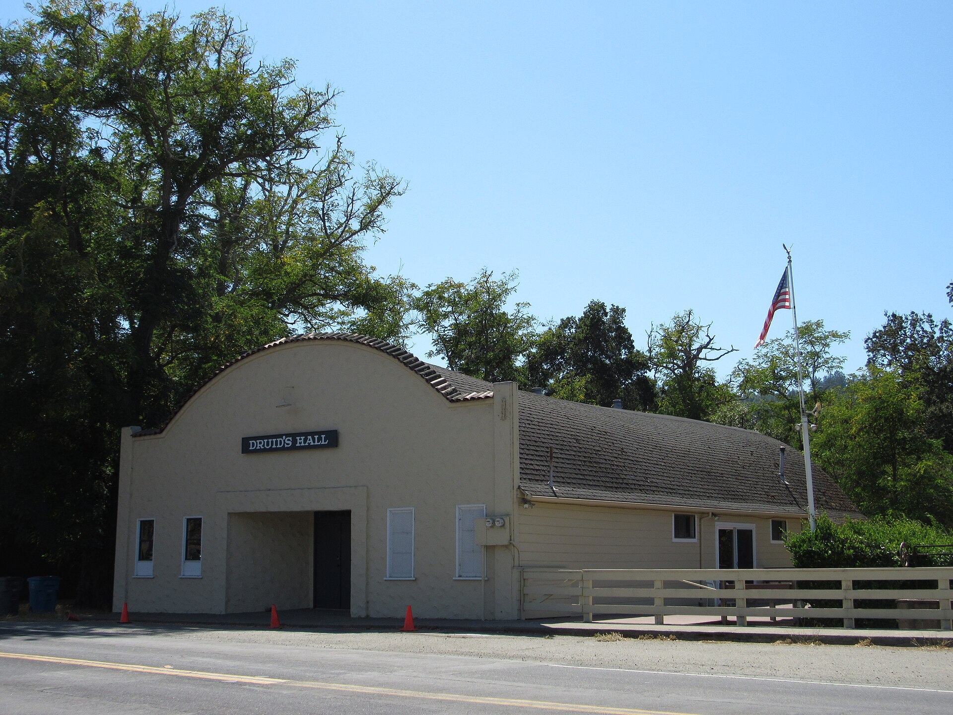 Druid's Hall in Nicasio, a stucco community hall with an arched entry, an American flag, and oak trees behind it.