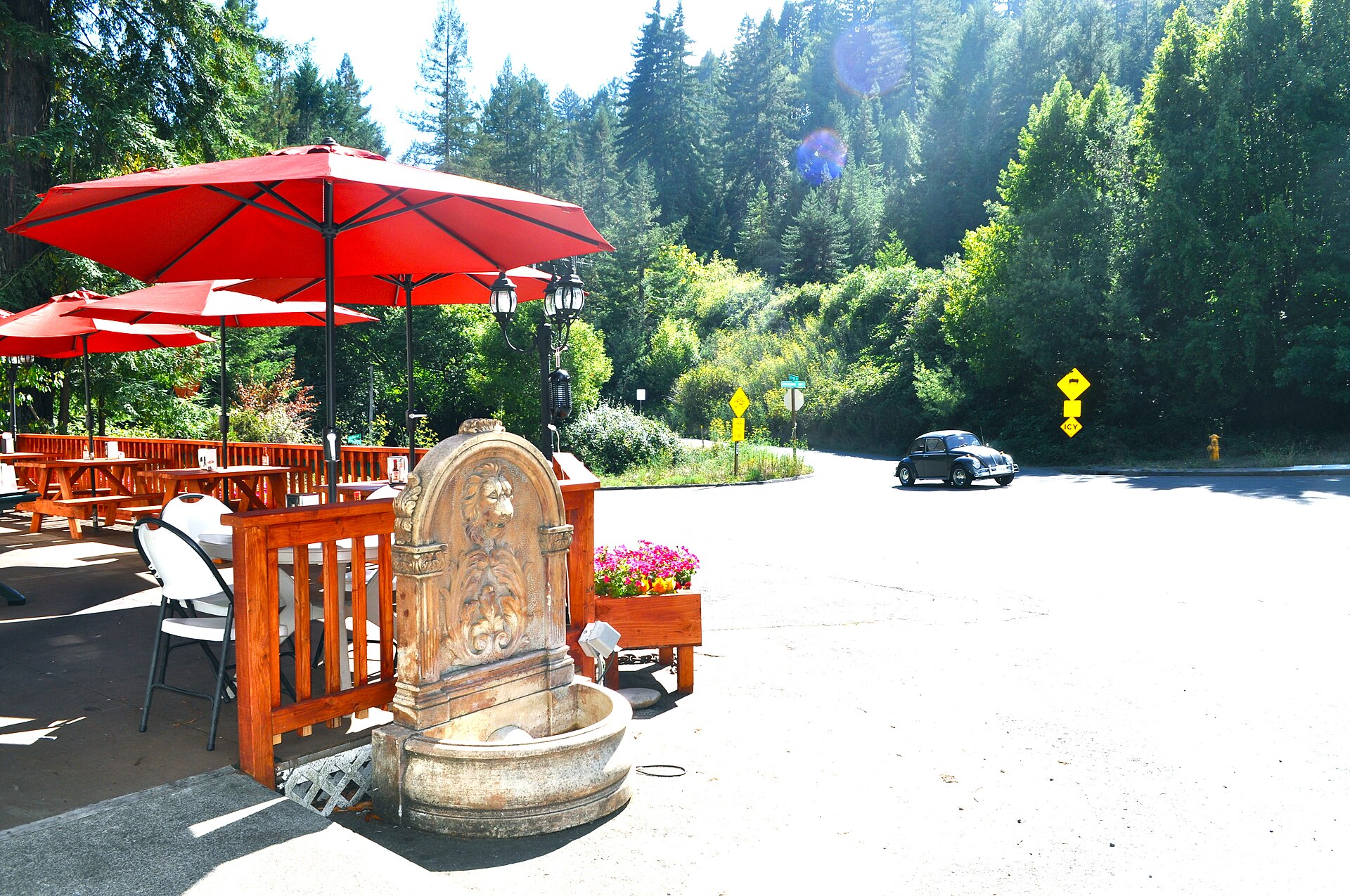An outdoor cafe terrace with red umbrellas and a stone fountain on a sunny street in Occidental.