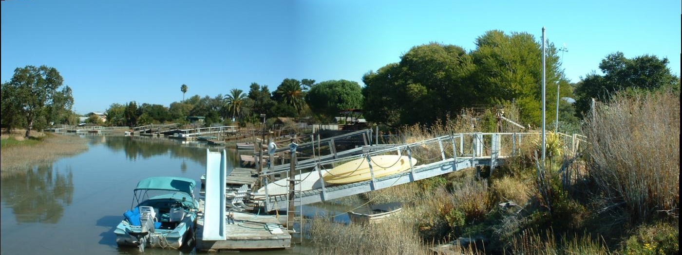 A canal in Santa Venetia with docked boats and a footbridge, lined with reeds and houses.