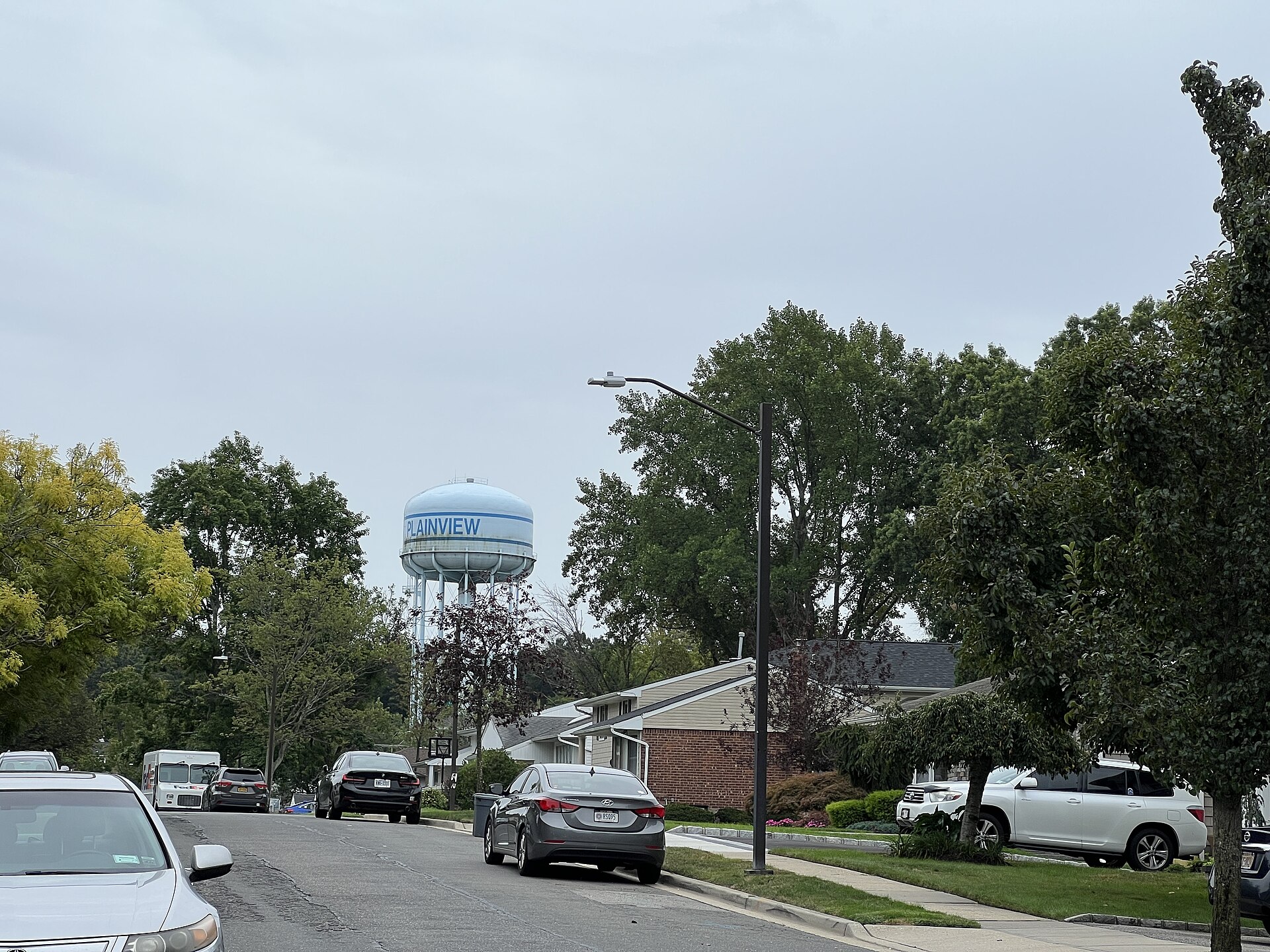A suburban street with parked cars, houses, and a water tower in the distance.