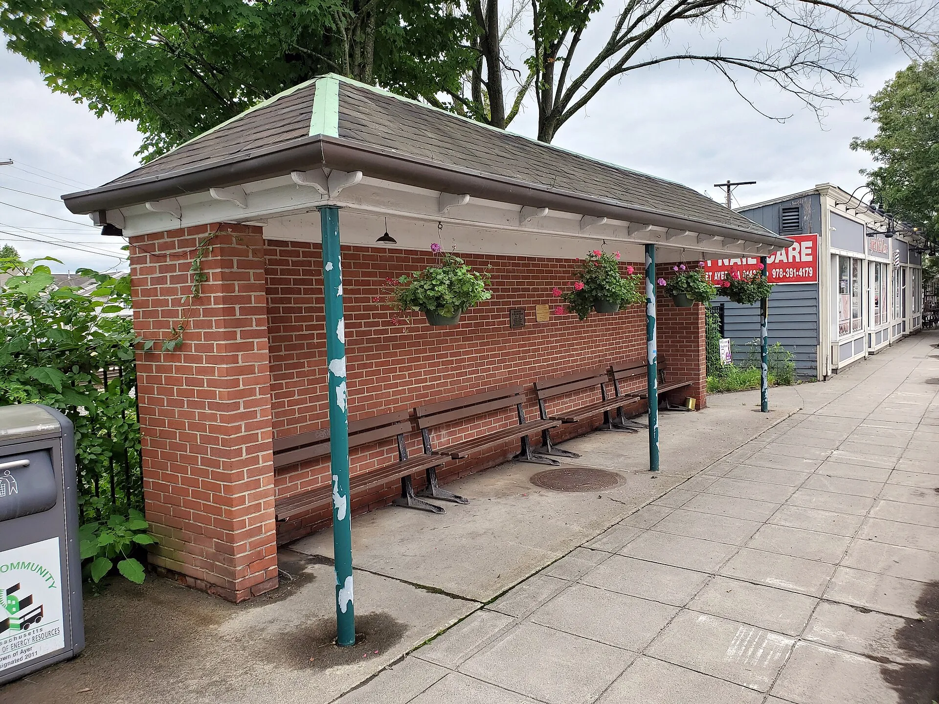 A small brick structure in downtown Ayer, seen in July 2021. It is likely a former streetcar and/or bus stop, and may be related to the under-track pedestrian tunnel once located at this point. Two plaques on the structure read: