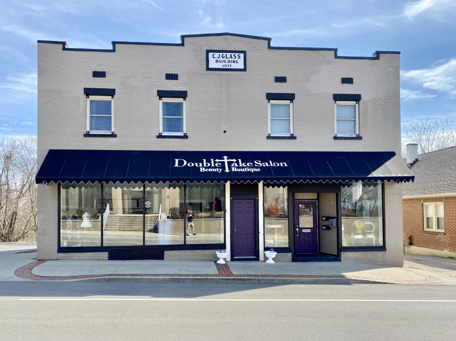 Built in 1937, this commercial building features a painted brick exterior, replacement windows, first floor storefronts, a stepped and gabled parapet with a datestone, and a concrete base.