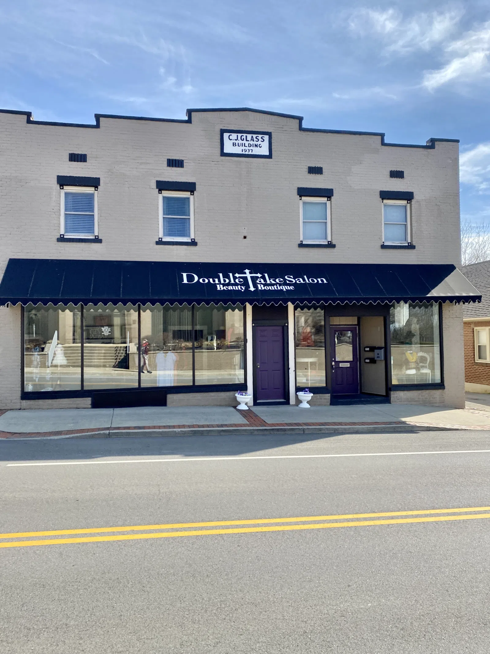 Built in 1937, this commercial building features a painted brick exterior, replacement windows, first floor storefronts, a stepped and gabled parapet with a datestone, and a concrete base.