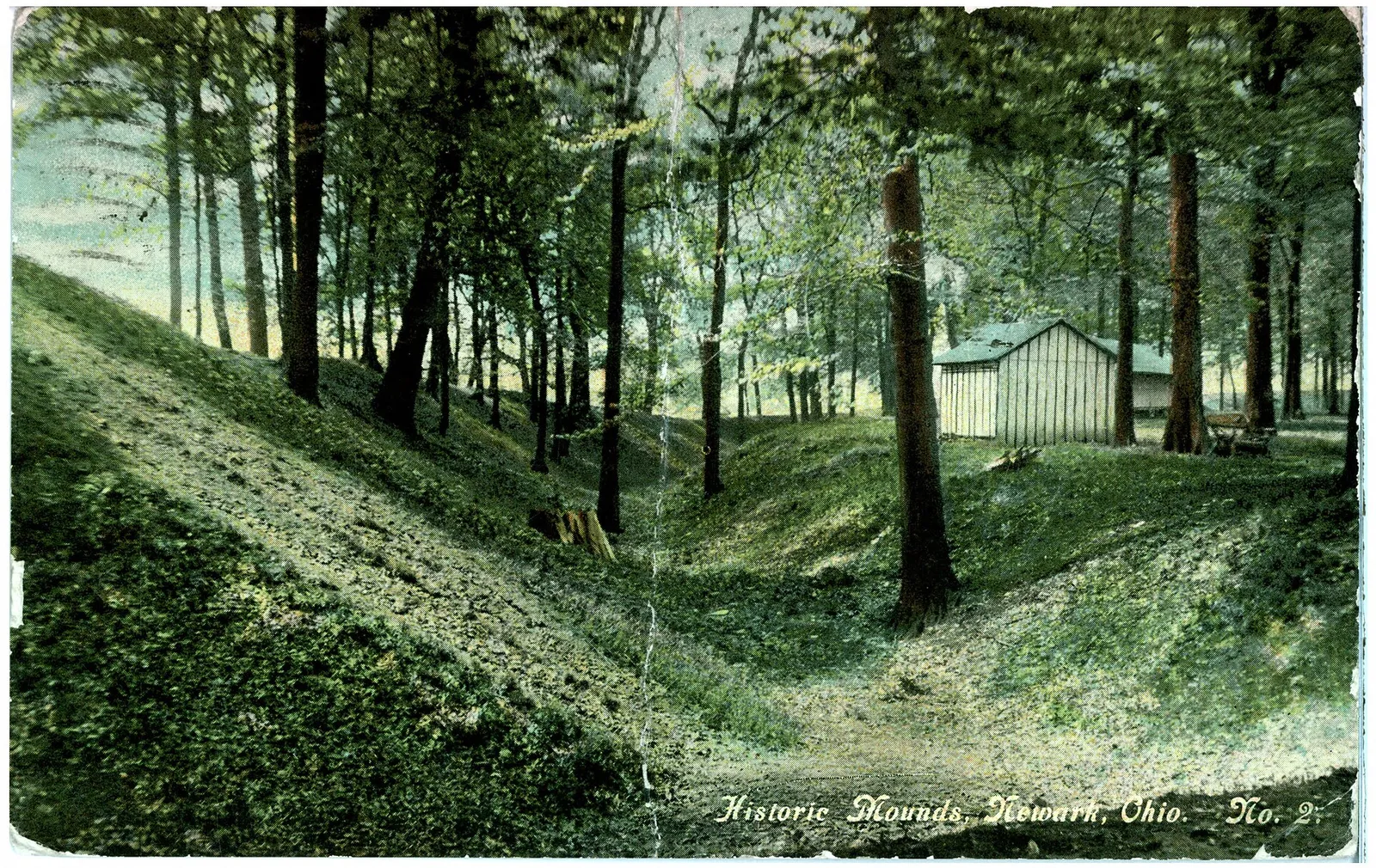 Colored photograph of tree covered mounds with buildings in background.