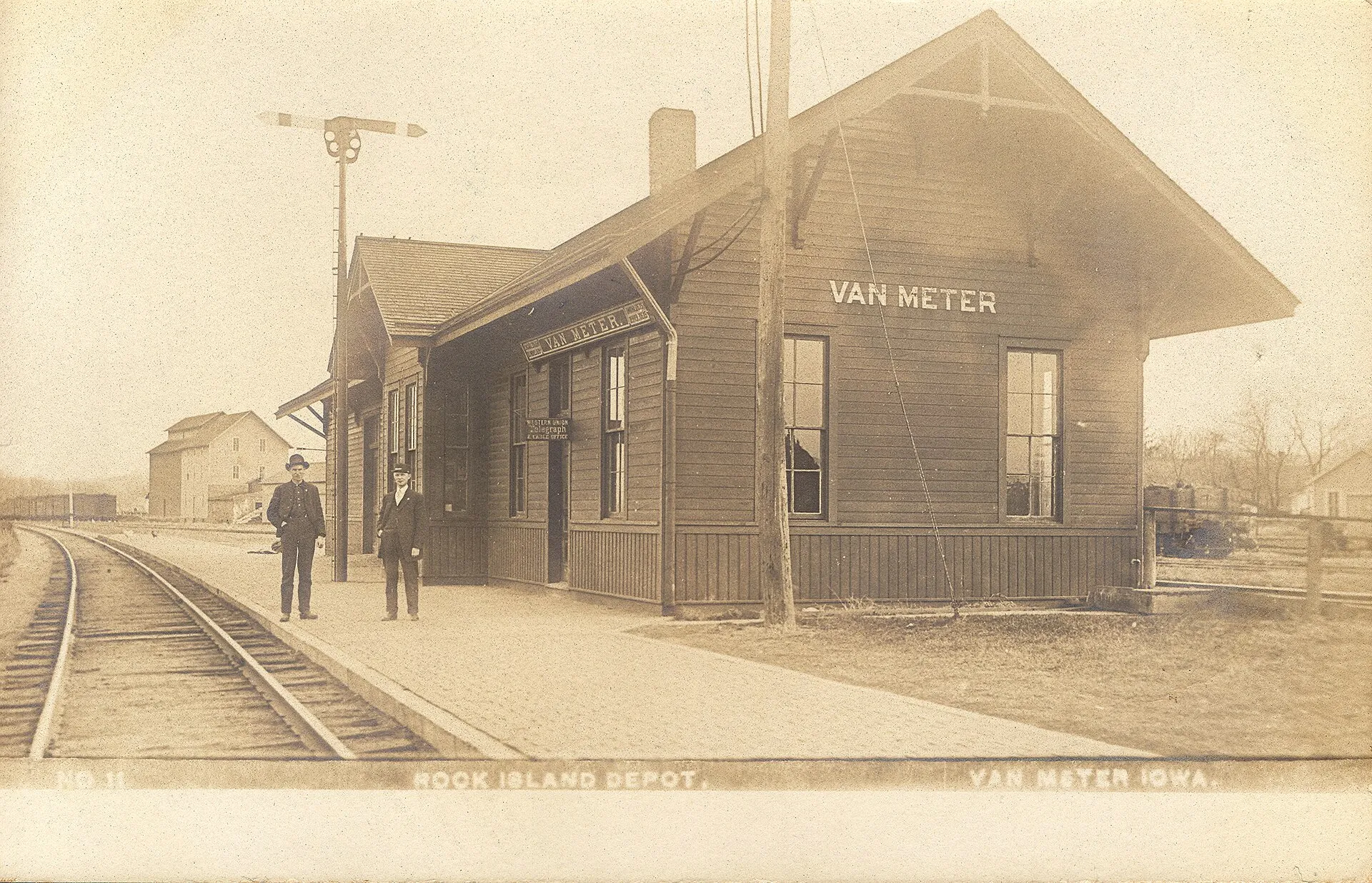 Two railroad employees are standing on a brick platform near a wooden depot with a train signal and telegraph office sign in the background. 1907 postmark. Number 11.