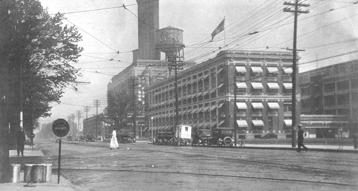 A woman walks across the street near the Ford Motor Company plant on Woodward Boulevard in Highland Park, Michigan around 1915. Terms associated with the photograph are: Woodward Boulevard (Highland Park, Michigan) | aut