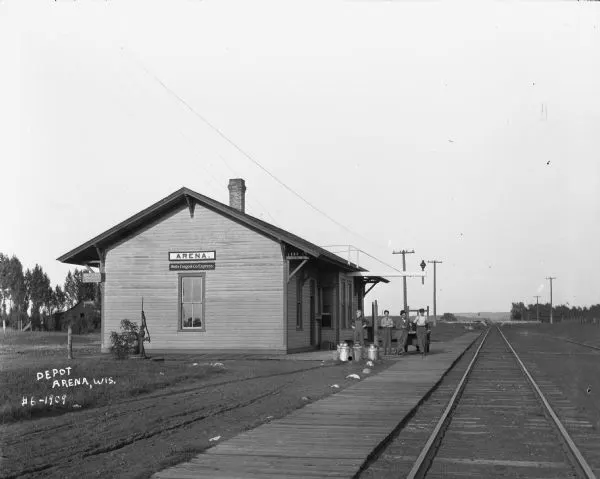 Exterior view from railroad tracks of the Depot. Four men are standing alongside the depot building near a wheeled cart. The building features two signs, one that says "ARENA," and the other that says "Wells Fargo & Co. Express." There is also a hand water pump on a landing near the depot on the lef