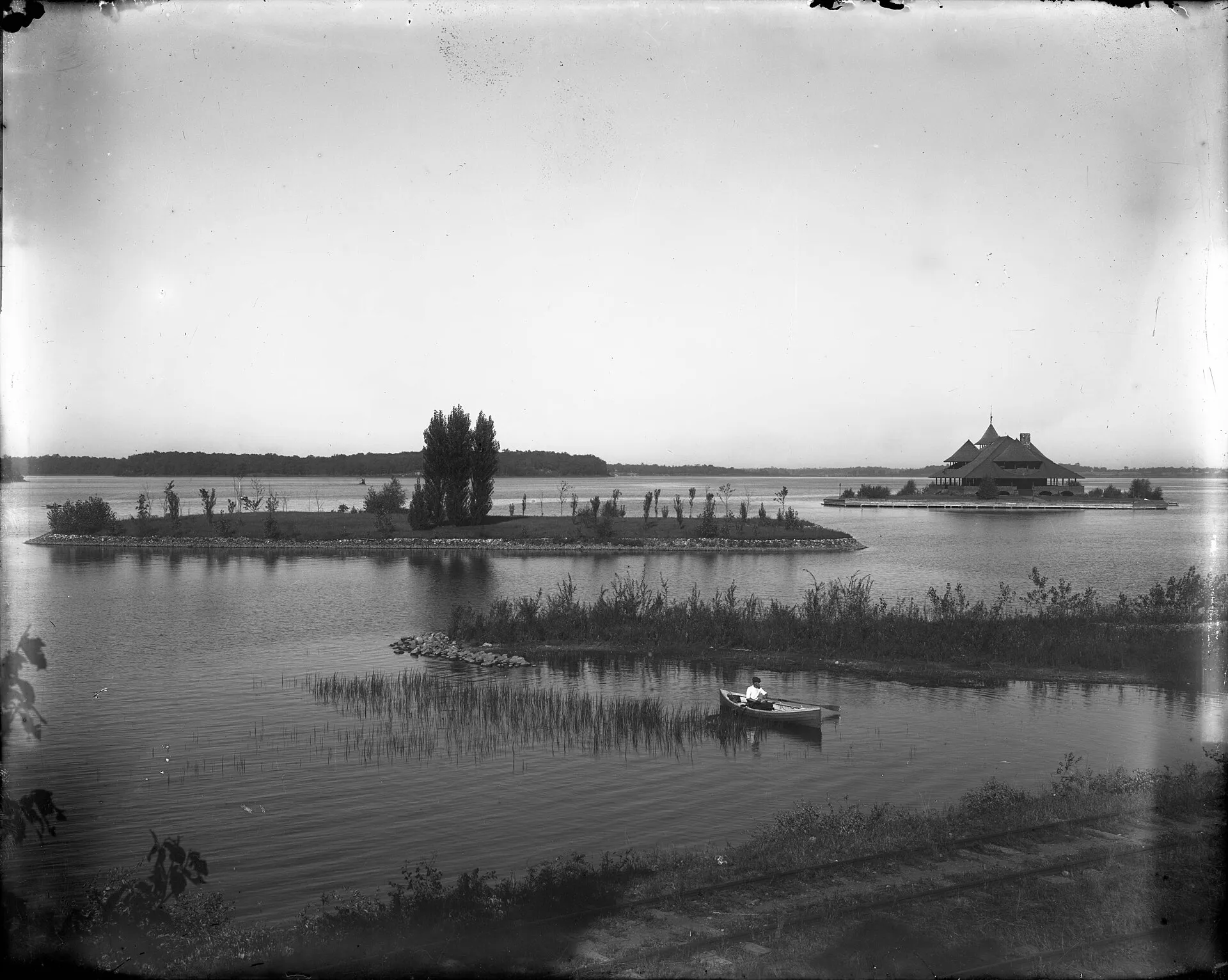Man is rowing in the foreground. Clubhouse is on an island. It has a very large gabled roof. BR0313.