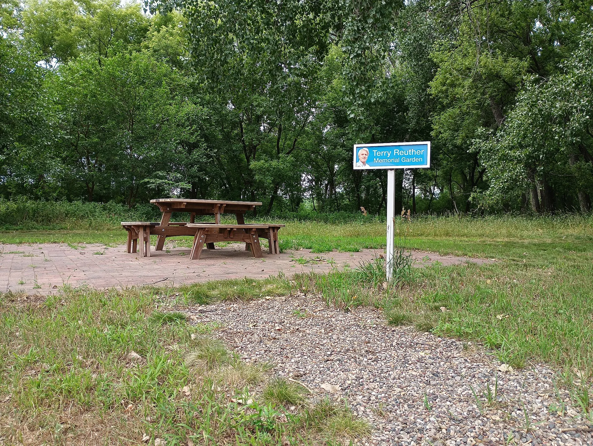 Terry Reuther (1944–2008) was a professor at Anoka-Ramsey Community College. The sign includes a color photo of him on a bright green background, akin to a street sign. The picnic bench has four attached benches. Photo taken by Ulysses Swanson..
