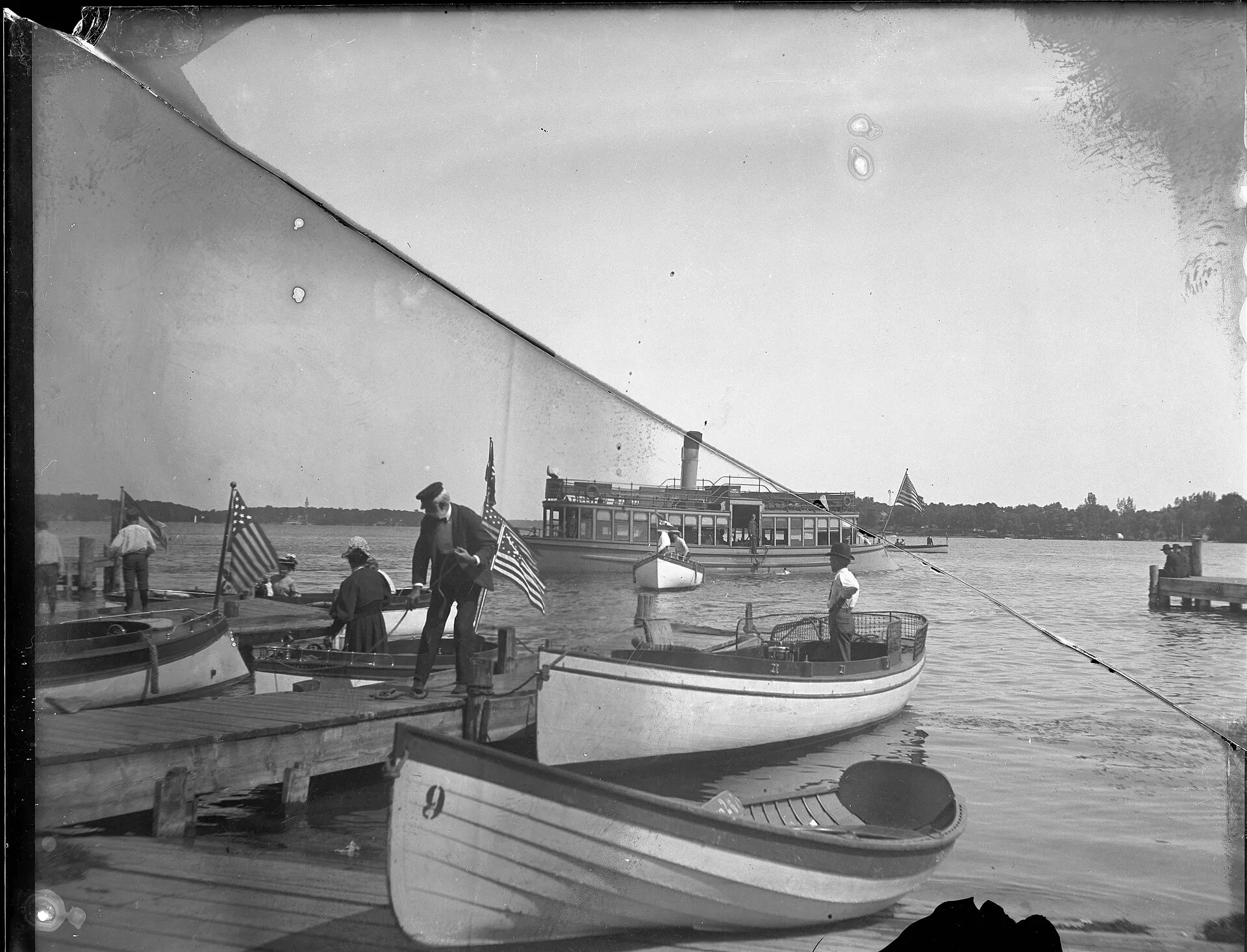 Launch is out a ways from the dock. Several people are tying up boats at the dock. The tower of the amusement park on Big Island is visible on the horizon at left. BR1016.