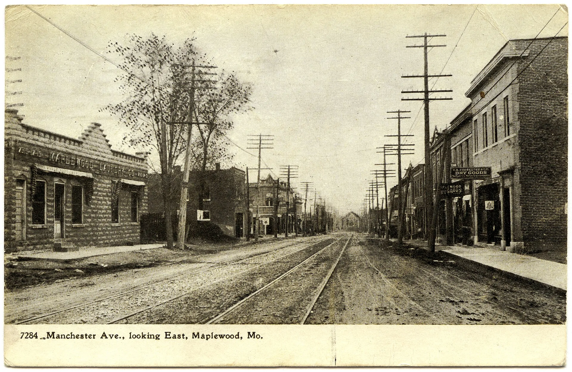 Horizontal, black and white postcard showing a photographic view looking east down Manchester Avenue at 7284 Manchester. Street car tracks run down the center of the dirt road, and businesses in one and two-story bric...